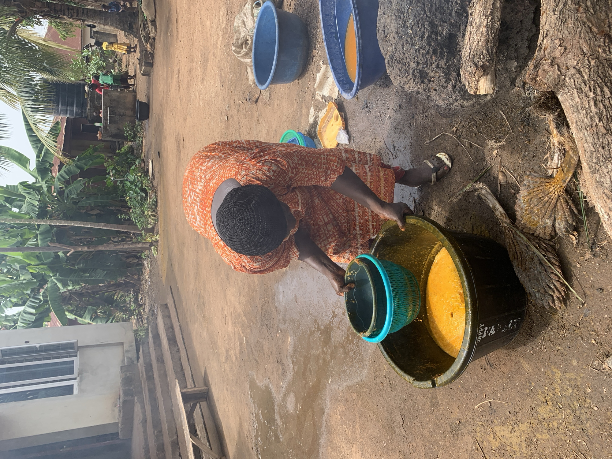 An overhead shot depicts a woman in Akwanga, Nigeria, wearing an orange patterned dress and a black head covering, bent over pouring liquid from a blue-rimmed green plastic basket into a black plastic bucket. The bucket contains a yellow liquid. Two blue plastic tubs, one filled with yellow liquid, sit nearby on the dusty ground. A large tree trunk is in the background, and children are visible in the distance near a water tower and a white building. The ground is muddy and damp.