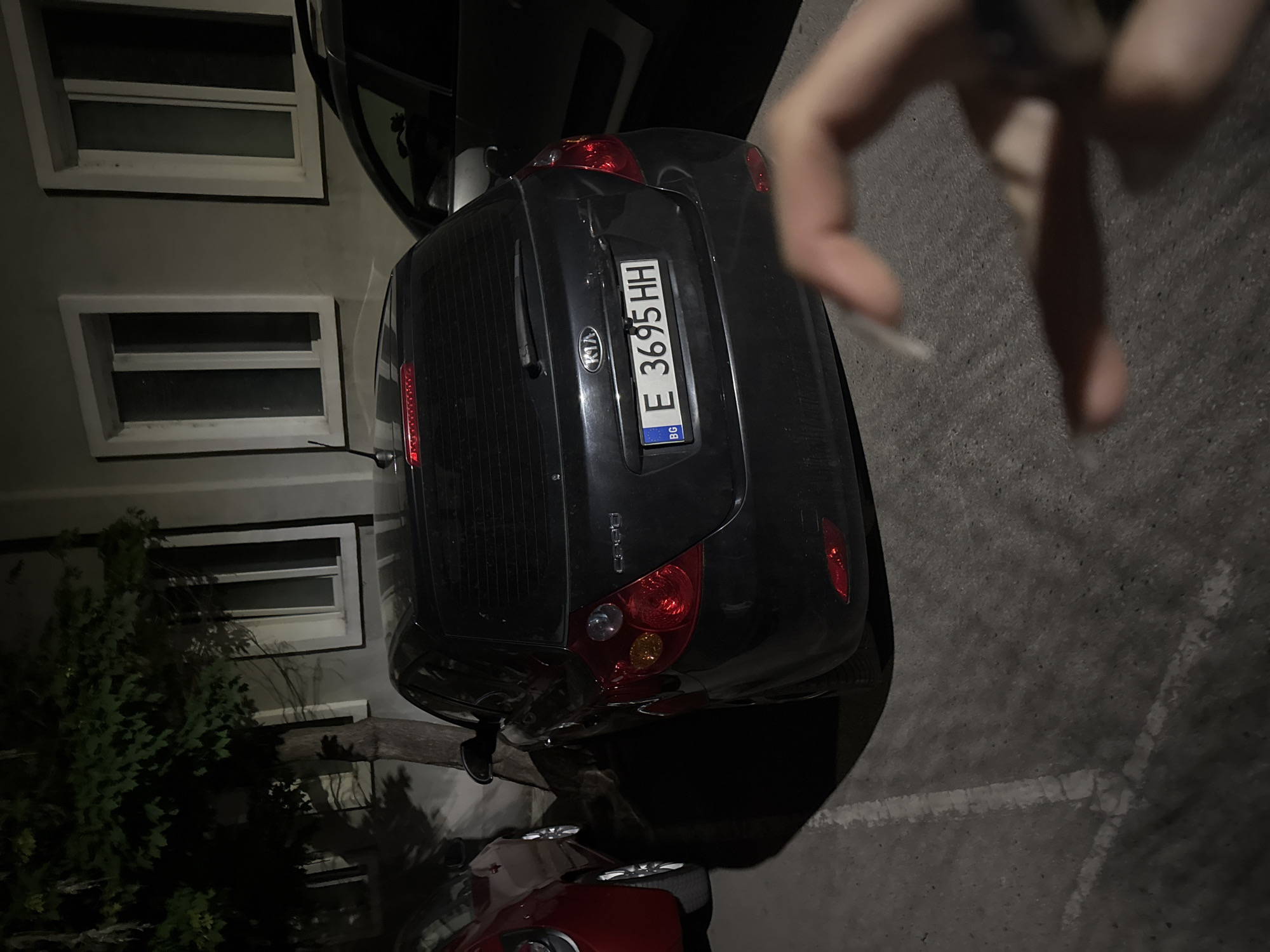 The image shows the rear of a dark gray Kia Ceed parked on a paved surface, likely asphalt, in an urban environment. The license plate number is E 3695 HH, indicating registration in Bulgaria. The license plate also shows "BG" and the Bulgarian flag. The car is parked next to a building with multiple windows. A tree is partially visible on the left side. The photo appears to be taken at night or in low light. The presence of the "Kia Ceed" logo on the rear of the car is visible. A partial view of another vehicle (likely red in color) can be seen beneath the Kia Ceed, in the lower part of the image. A person's hand is blurred and partially visible in the upper right. There are painted parking lines on the ground. The location appears to be Blagoevgrad, Bulgaria, based on the Bulgarian license plate.
