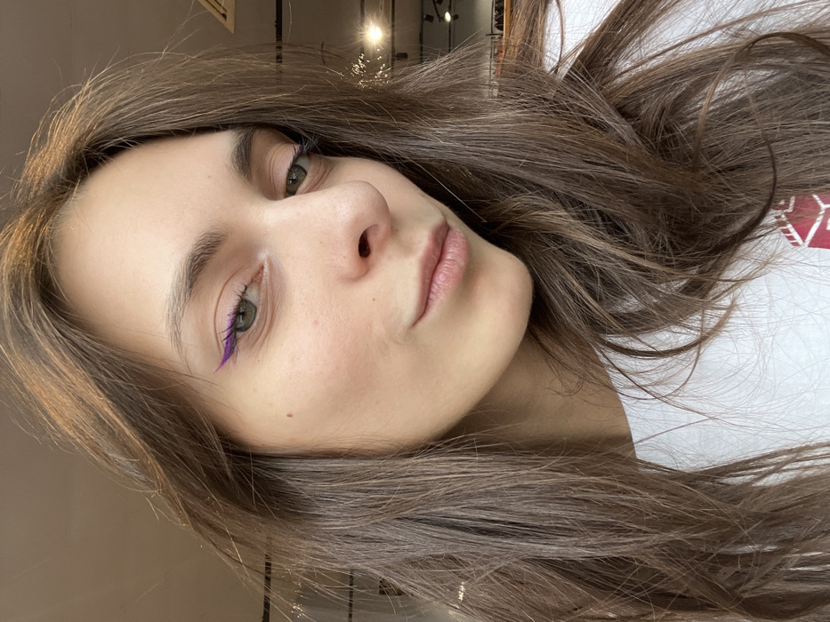 A close-up, chest-up portrait features a young adult female indoors, looking directly at the camera. She has long, wavy brown hair flowing over her shoulders. Her eyes, appearing green or hazel, are distinctly accented with prominent, winged purple eyeliner on the upper lash lines. Her eyebrows are natural and full, and her lips are closed in a subtle, relaxed smile. A small mole is visible on her left cheekbone. She wears a white top, with a partial red graphic design visible on the lower right portion of the garment. The background is significantly blurred, indicating an indoor commercial or public setting. Visible elements in the background include overhead track lighting fixtures, a ceiling-mounted ventilation unit, and indistinct shapes possibly suggesting shelving or displays. The specific location within the city of Krasnoyarsk, Russia, is not discernible from the visual information presented in the image.