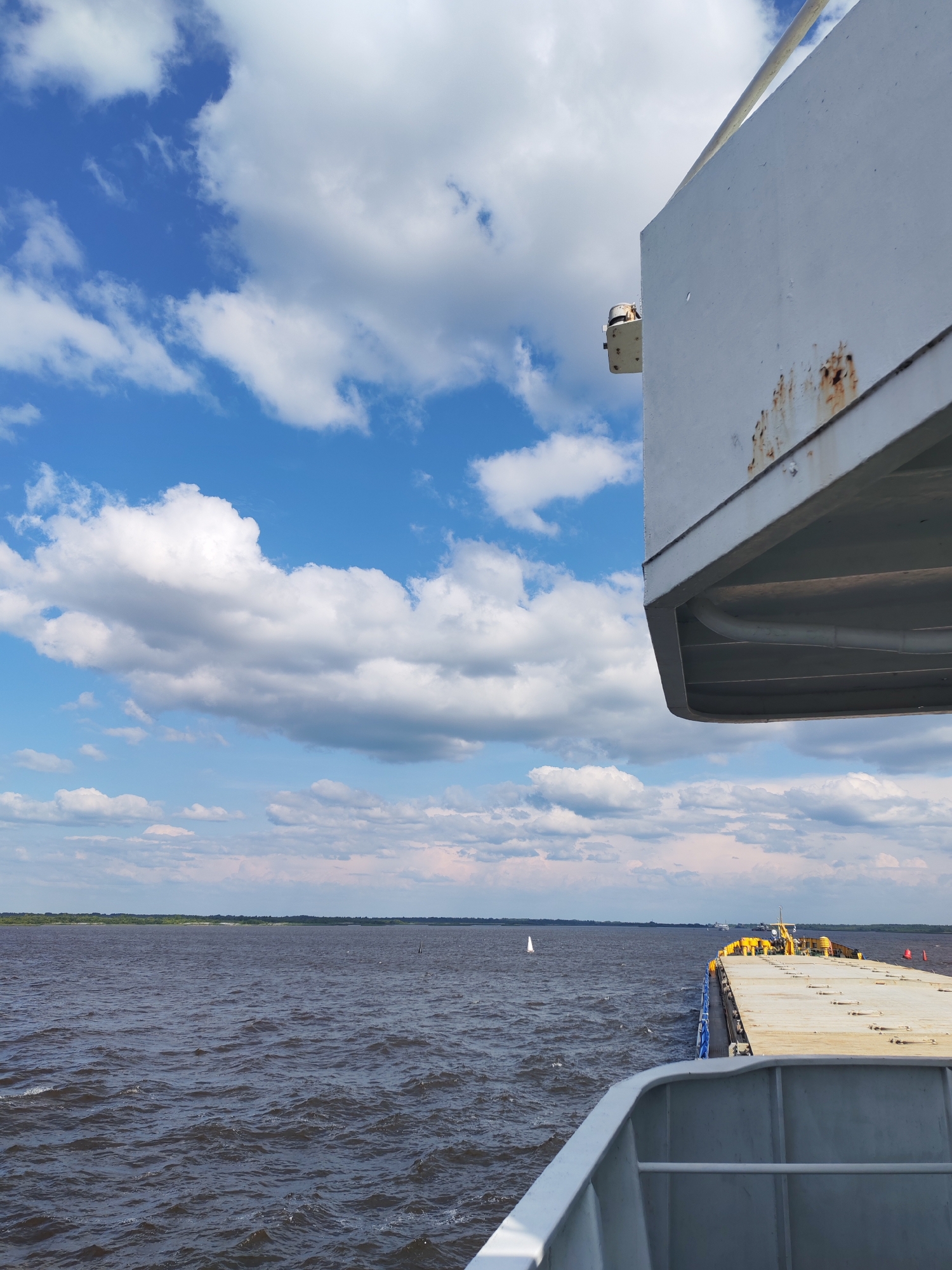 The image captures a view from a vessel on a wide body of water, likely a river, under a sky filled with scattered white clouds against a bright blue backdrop. In the foreground, a portion of a ship's deck and superstructure is visible on the right side, showing a weathered white surface with streaks of rust. The water is dark and choppy, suggesting a moderate breeze. In the distance, a thin strip of green land borders the horizon, indicating a coastline. A small white sailboat can be seen on the water in the mid-ground. Other vessels are faintly visible on the horizon. The scene is a static, outdoor environment.