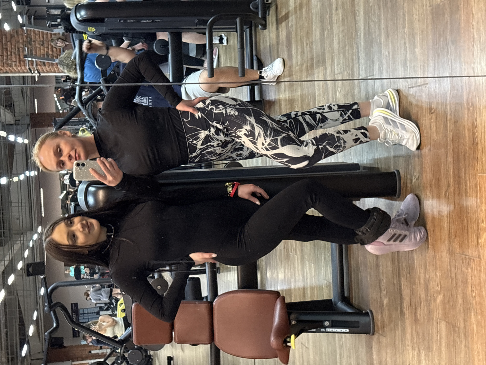 The image, currently rotated 90 degrees clockwise, depicts two women posing for a selfie in a gym, as seen through a large mirror reflection.
**Subjects:** The primary subjects are two women in athletic attire. The woman on the left has blonde hair, light-colored eyes, and holds a smartphone with a glittering case, actively taking the photo. The woman on the right has dark hair and a gentle smile. Several other indistinct individuals are visible in the background reflections, presumably also exercising.
**Scene:** The setting is an indoor gym or fitness center. The floor is made of light-colored wooden planks. Various exercise machines with black frames and brown-padded seats are visible throughout the space. Overhead lighting fixtures provide illumination. Exposed brickwork can be seen in the reflected background, adding an industrial touch to the gym's decor.
**Activity or event:** The two women are captured in the act of taking a selfie together during their time at the gym. They appear to be friends or workout partners. The overall scene suggests a routine gym visit.
**Notable details:**
The blonde woman wears a black long-sleeved top and black leggings with a striking white abstract pattern, paired with white athletic shoes featuring yellow accents. The dark-haired woman is dressed in a plain black long-sleeved top and black leggings, complemented by light pink athletic shoes with black stripes and a subtle black choker necklace. She also wears a red and gold bracelet on her right wrist. The image is clearly a mirror shot, creating a sense of depth and reflection of the gym's interior.
**Visible text:** The brand "ADIDAS" and its distinctive three stripes are visible on the light pink athletic shoes worn by the dark-haired woman. The three stripes are also visible on the white shoes of the blonde woman. Partially obscured and reversed Cyrillic text, appearing as "РТА," is faintly visible on a blue garment worn by a background figure.