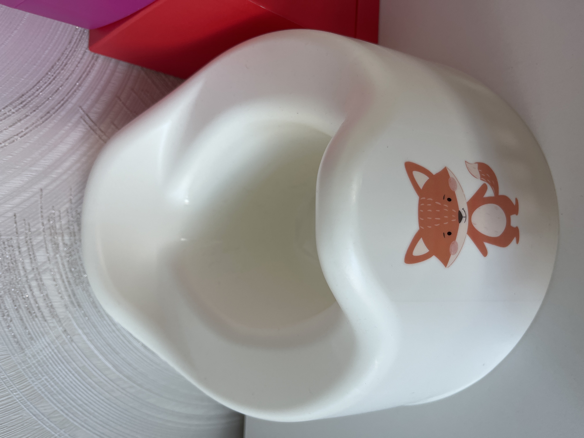 The image displays a white plastic potty training seat, designed for children. The potty is the central and most prominent object, oriented as if viewed from the front-right side. It features a curved, ergonomic design with a raised backrest and a small splash guard at the front.
A notable detail is the cartoon illustration of a fox on the right side of the potty's exterior. The fox is depicted with an orange body, a white belly, white inner ears, and a white-tipped tail. It has small black eyes and a reddish-pink nose or cheek blush. Its posture appears playful, with one front paw slightly raised.
In the background, behind the potty, several other objects are partially visible. To the upper left, there is a red, rectangular object, possibly a storage box or part of a larger piece of furniture. Above this red object, a sliver of a purple or magenta item can be seen. The wall behind these objects and the potty is light-colored, possibly white or light grey, and features a textured, wavy pattern that suggests wallpaper or a decorative wall finish.
The lighting is bright and even, indicating an indoor setting. There are no people present, and no specific activity is taking place. No visible text is present on the potty or in the background. The overall scene suggests a functional item within a child-friendly or domestic environment.