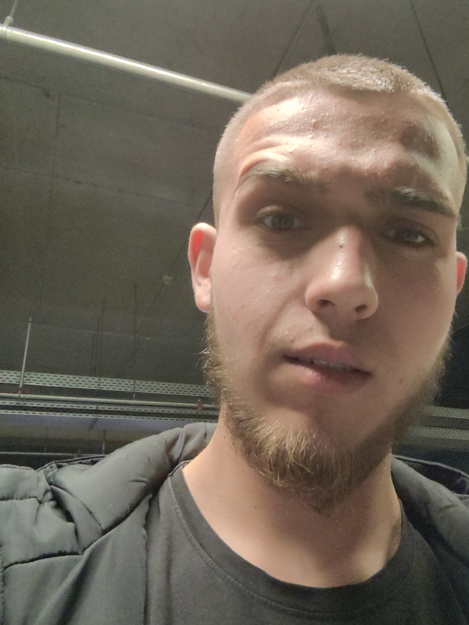A selfie of a young man with a closely shaved haircut and a beard. He has a scar on his forehead. He is wearing a black t-shirt and a black jacket. The background appears to be a concrete ceiling with exposed pipes and cable trays, possibly in an underground parking garage. The location could be Sofia, Bulgaria, based on the image context.