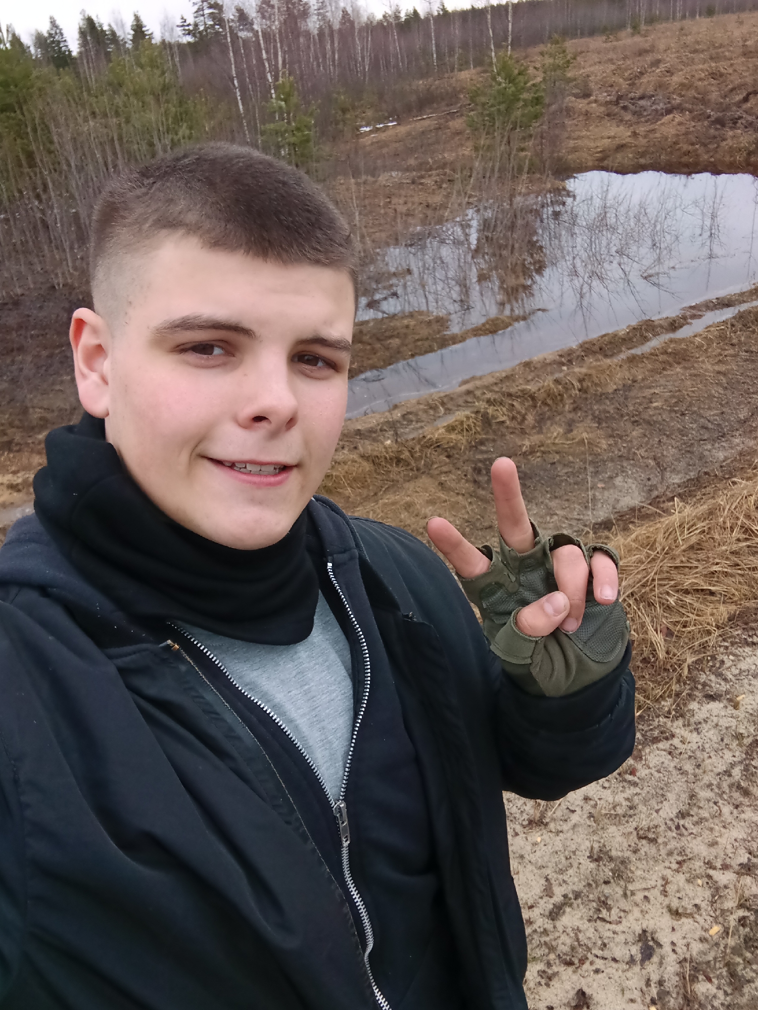 A selfie of a young man with short, light-brown hair is taken outdoors. He is wearing braces, a black jacket, a gray shirt, a black neck warmer, and green fingerless gloves. He is giving a peace sign with his gloved hand.
The background shows a body of water, likely a swamp or large puddle, reflecting trees and the overcast sky. The terrain is brown and muddy, with dry grasses visible. In the distance, there are bare trees and some evergreens.
Based on the information provided, this photo was taken in Krasnyi Mayak, Russia. The specific location within the city is not identifiable from the image.