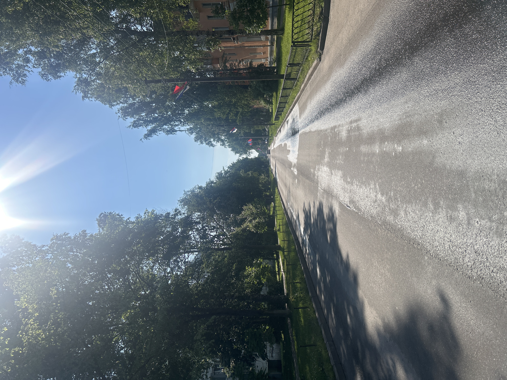The image depicts a straight, paved road lined with mature trees on both sides. The road surface appears somewhat wet and reflective, suggesting recent rain or a street cleaning. On the right side of the road, there is a black metal fence bordering a grassy area and buildings. Several flags, red and white, are visible on poles along the street. The sky is bright blue with a strong sun, causing lens flare in the upper left corner. The scene is likely a residential or public street in Posyolok Imeni Morozova, Russia, captured on a sunny day. There are no people or vehicles clearly visible in the immediate foreground, though a vehicle can be seen in the distance down the road.