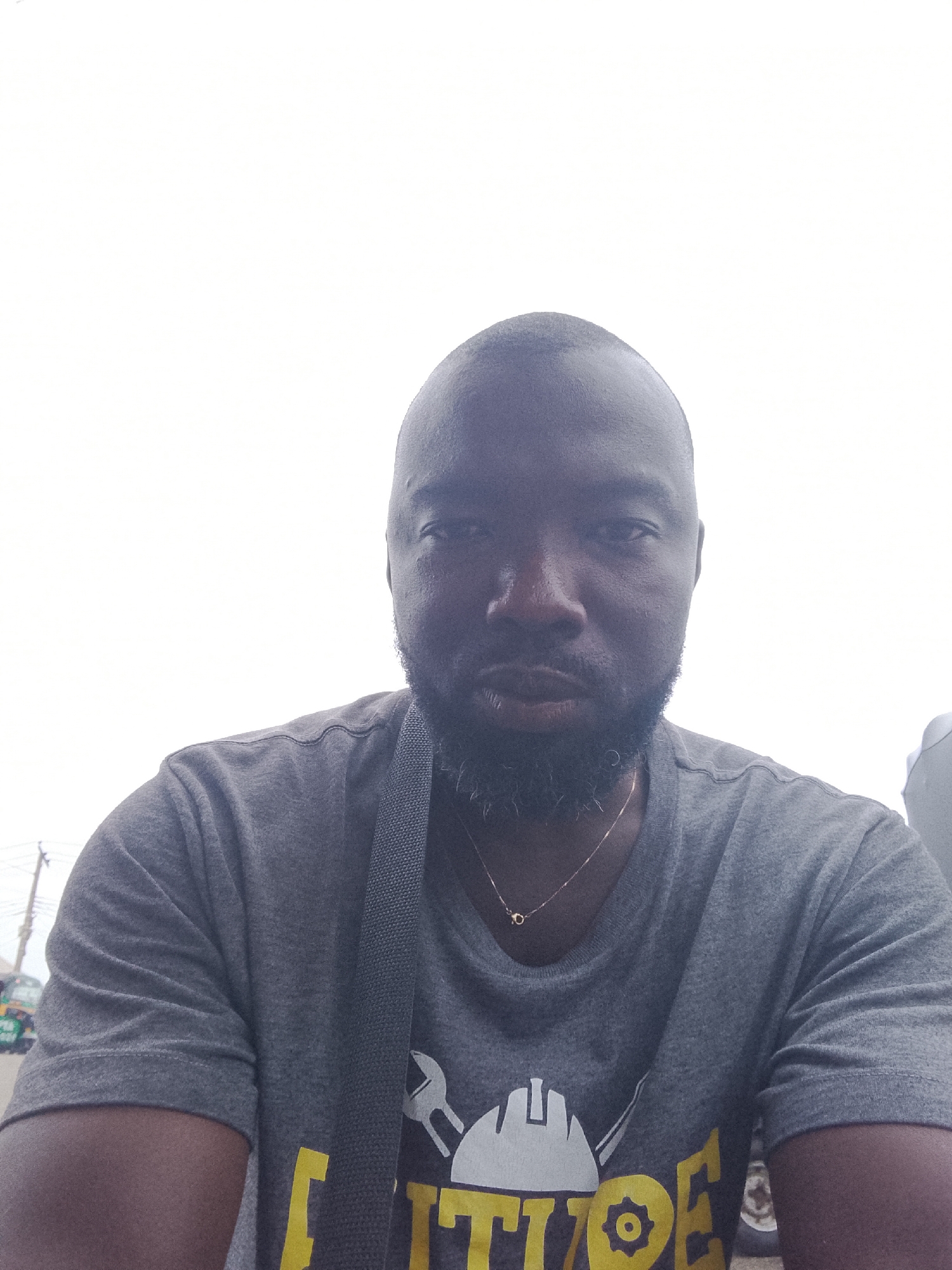 A close-up portrait of a dark-skinned man with a shaved head and a full beard. He is wearing a gray t-shirt with a graphic design and a thin gold chain necklace. A gray strap is visible across his chest. The background is bright white and overexposed, suggesting it is outdoors. Based on the prompt, the location is likely in the city of Gwong, Nigeria, although no specific landmarks are visible.