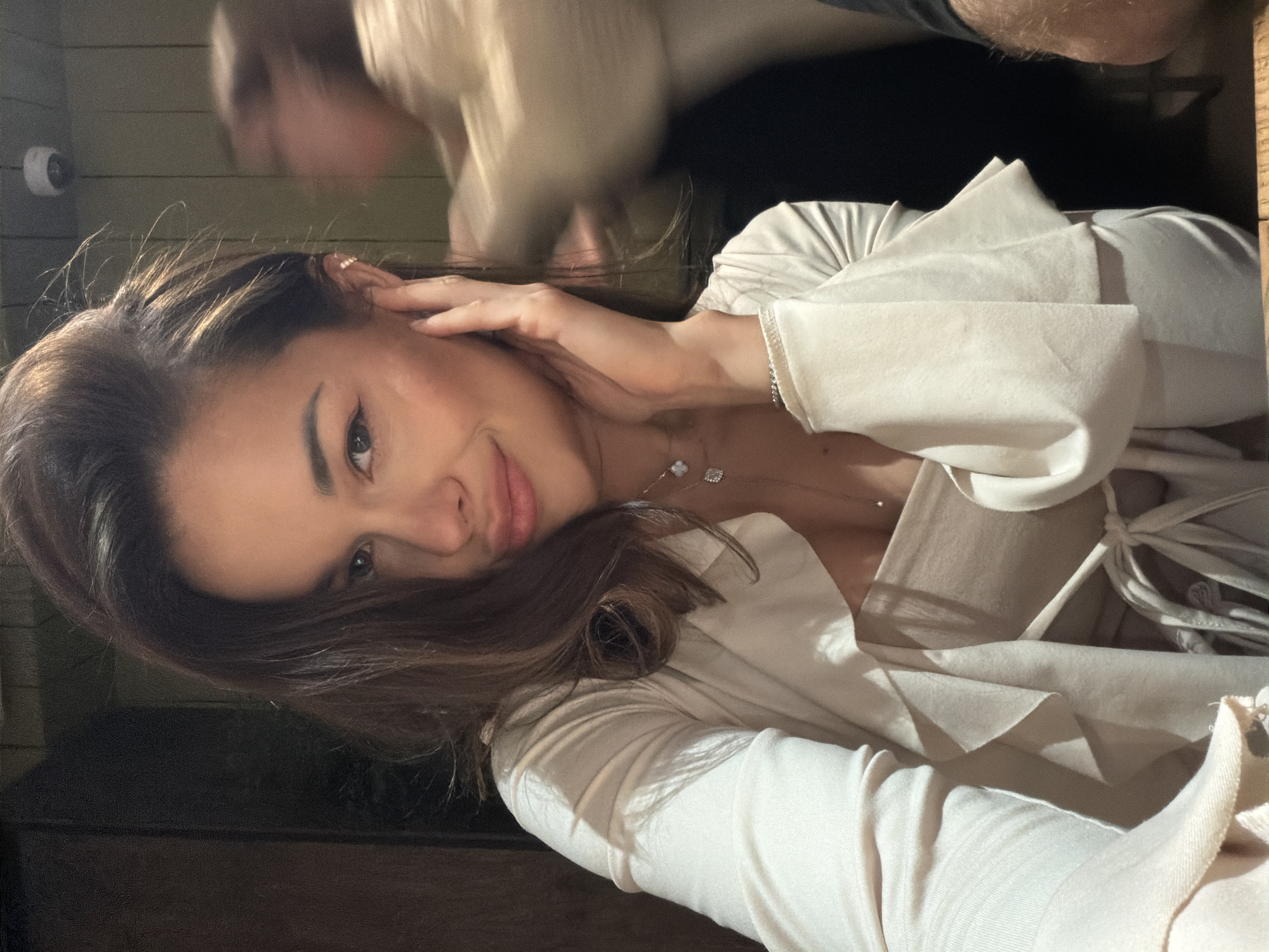 A woman with long brown hair, wearing a cream-colored blouse and necklace, is touching her face in a room. In the background, a blurred figure is visible, and there appears to be a security camera mounted on a wall. The location is difficult to determine as the image does not contain location-specific clues. Based on the information provided, there is no definitive way to identify whether this scene is in the city of Krasnoyarsk, Russia.