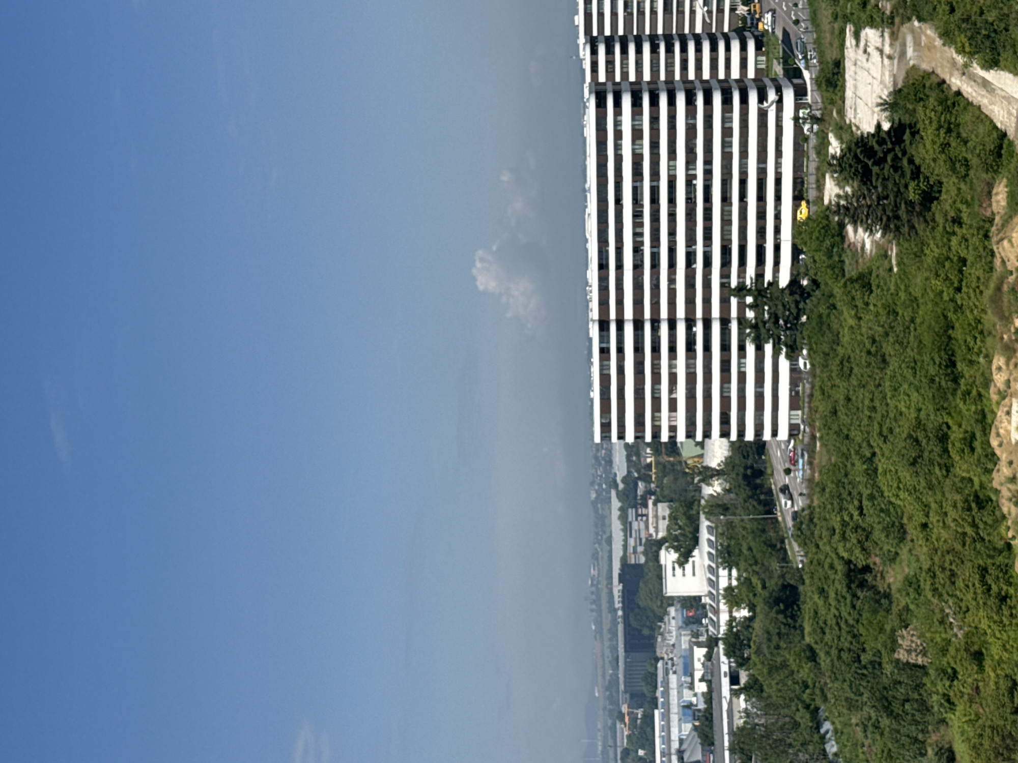 This image presents a panoramic view of an urban landscape under a clear, bright blue sky, with a few faint wisps of clouds. The scene appears to be captured from an elevated perspective, looking down and across a city.
On the right side of the frame, several modern, multi-story buildings dominate, characterized by a striking design of alternating vertical white and dark (possibly dark glass or shadowed) stripes. These buildings appear to be part of a larger complex, with at least two prominent structures visible closer to the foreground.
Adjacent to these buildings and extending along the right and bottom-right portion of the image is a steep, heavily vegetated hillside or cliff. This natural incline is covered with dense green trees and bushes, and sections of exposed rock or concrete retaining walls are visible, suggesting stabilization or landscaping efforts. Roads wind through the urban area at the base of these buildings and alongside the hillside, with several vehicles, including cars and possibly larger trucks or buses, discernible on them.
In the mid-ground and extending into the distance on the left and bottom-center, a sprawling city is visible, with numerous buildings of varying sizes and styles stretching towards the horizon. The distant elements are slightly obscured by atmospheric haze, indicating a potentially wide-ranging view. The overall impression is a daytime scene, possibly during good weather, highlighting a blend of contemporary architecture with natural topography within an urban setting, likely in Varna, Bulgaria. No visible text is clearly legible in the image.
