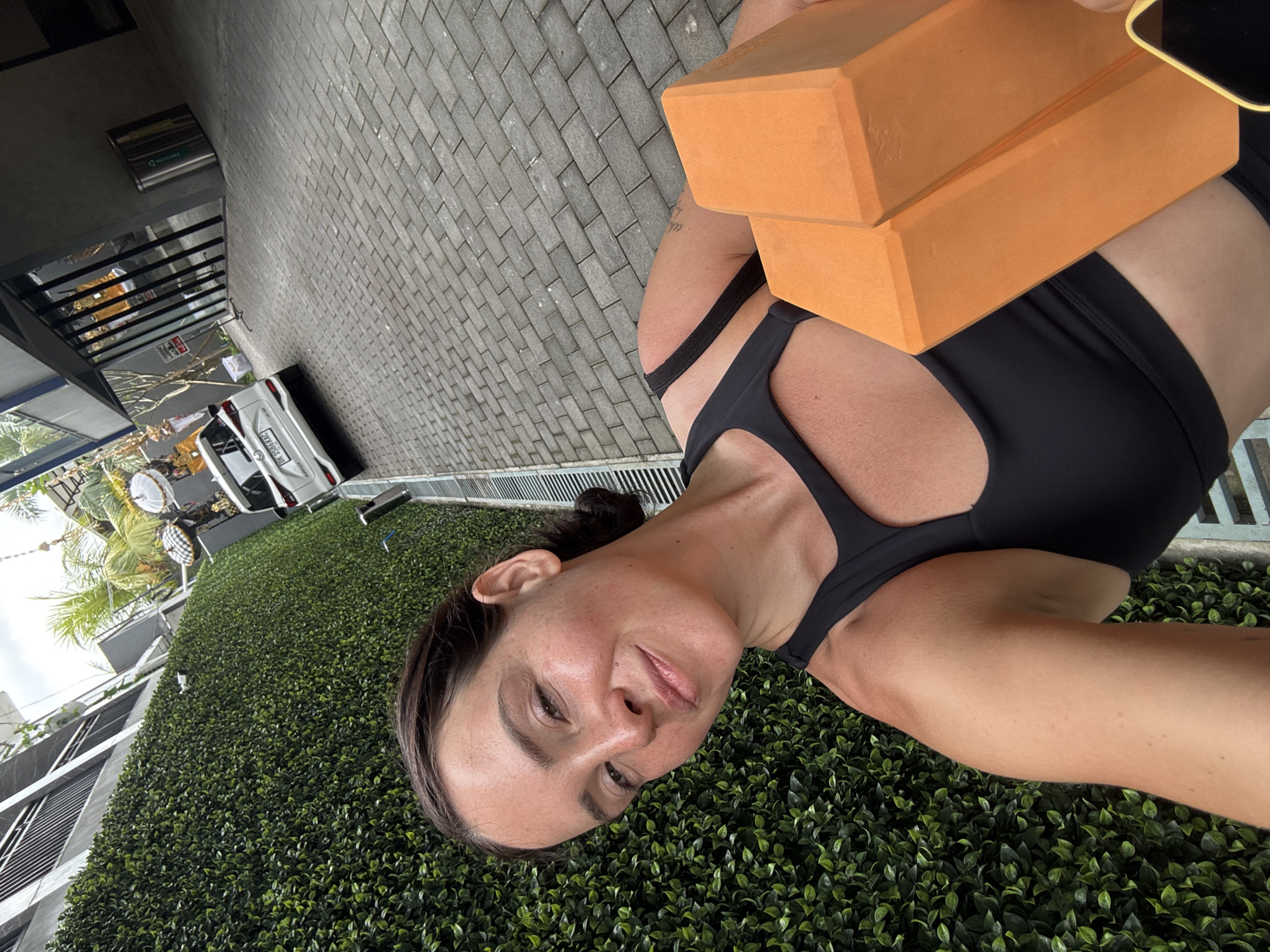 A woman with dark hair pulled back stands in front of a green hedge wall, holding two orange yoga blocks. She is wearing a black sports bra. In the background, a white car is parked on a paved driveway. Buildings and palm trees are also visible. The scene appears to be in North Kuta, Indonesia.