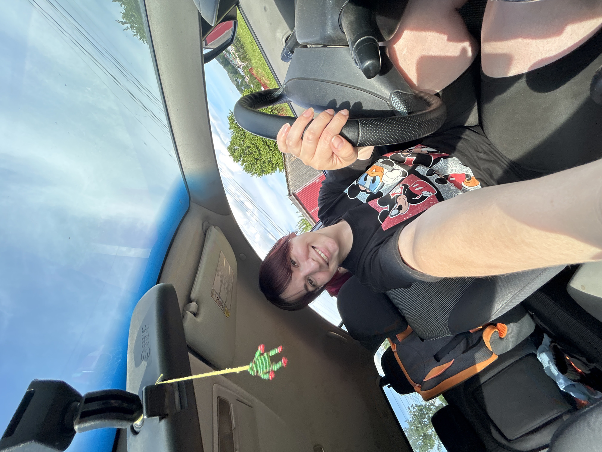 A young woman with dark hair and bangs, wearing a black t-shirt with a Mickey Mouse graphic, is sitting in the driver's seat of a car, holding the steering wheel. She is smiling and looking at the camera, with her arm extended as if taking a selfie. The interior of the car is visible, including the dashboard, rearview mirror with a green frog-shaped air freshener hanging from it, and a child car seat in the back. The exterior view through the windshield shows a sunny day with a blue sky, some trees, and buildings. The car appears to be parked on a street in the city of Fokino, Russia, based on the context.