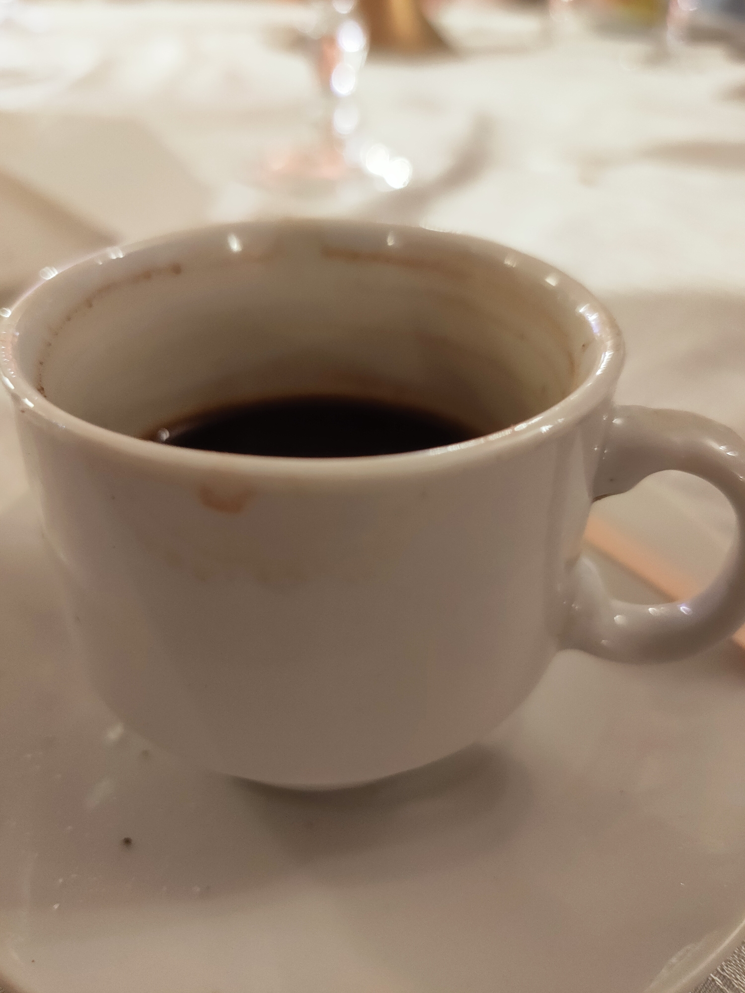A close-up, slightly blurry image shows a white ceramic coffee cup filled with dark coffee. The cup has a stain of coffee residue on the outside, near the rim. The cup is sitting on a white saucer, which is on a textured surface, possibly a tablecloth. In the background, out of focus, are hints of a light-colored tablecloth, some glassware, and possibly other objects on a table. The lighting appears to be indoor ambient light. The image is taken from a slightly elevated angle, looking down at the cup. There is no discernible information about the location in the city of Mornag, Tunisia, in this image.