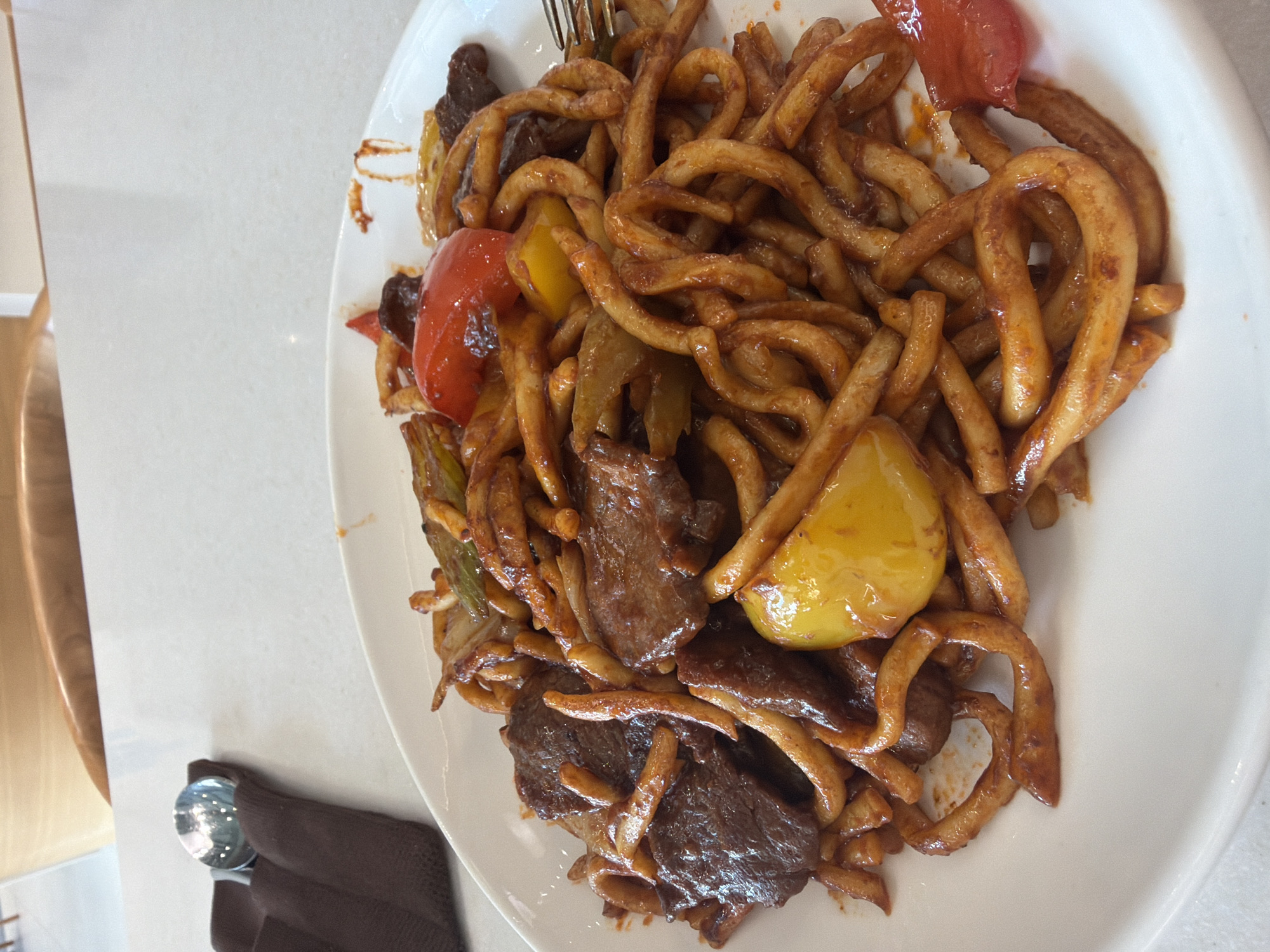 A close-up, overhead shot captures a plate of food, likely noodles with beef and vegetables, on a white surface. The dish features thick, golden-brown noodles coated in a glossy, reddish-brown sauce. Interspersed among the noodles are slices of dark, cooked beef and pieces of colorful bell peppers in shades of red and yellow, along with some green vegetables. A fork is visible on the right side of the plate, partially obscured by the food. In the upper left corner, a brown napkin is folded, with a silver spoon peeking out from under it. The background shows a blurred view of a light-colored table and wooden chairs, suggesting a restaurant setting. The overall impression is of a hearty and flavorful meal. This image appears to have been taken at a restaurant in Astana, Kazakhstan.