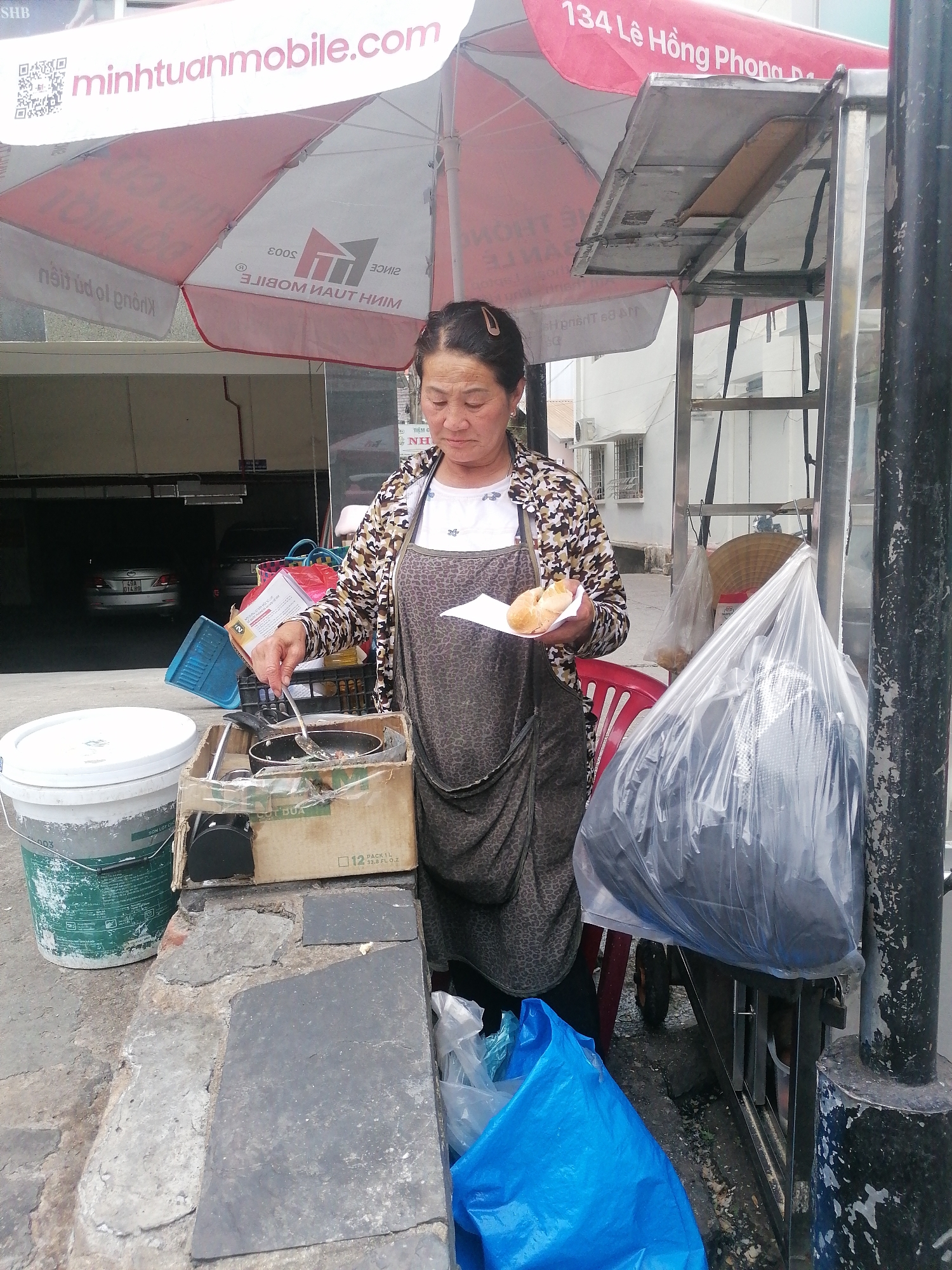 A street vendor is cooking outdoors in Da Lat, Vietnam, under an umbrella. The woman, with her hair pulled back, wears a patterned jacket, a dark apron, and a white shirt. She holds a plate with fried items in her left hand and uses a metal utensil in her right to cook in a small pan set atop a cardboard box. Next to her cooking setup is a white bucket and a concrete block. Behind her, a red plastic chair is partially visible, along with a cart covered by a large plastic bag. The umbrella overhead features the name "minhtuanmobile.com" and "134 Lê Hồng Phong". In the background, cars are parked in a garage.