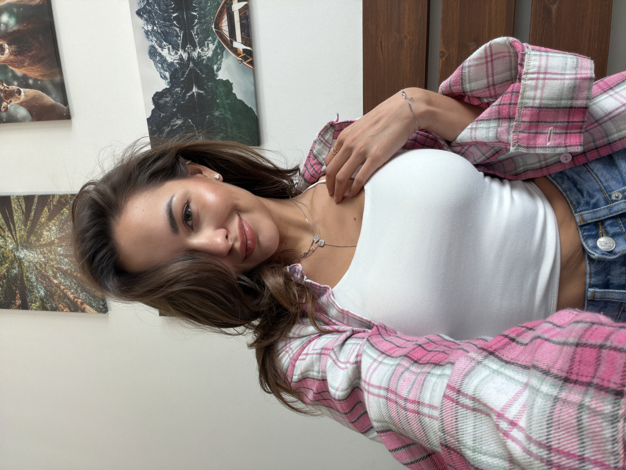 A young woman with long, dark brown hair, a slight smile, and a friendly expression is captured in an indoor setting, likely in Krasnoyarsk, Russia. She is lying down or leaning back, looking directly at the camera with her body oriented horizontally in the frame.
She is wearing a white sleeveless top, layered with an open, long-sleeved plaid shirt featuring shades of pink, white, and light green. Her lower attire includes blue denim jeans, on which a silver button with the embossed text "TRUE RELIGION" is visible near the waist. Accessories include a delicate silver necklace with two small clover-shaped charms, a silver stud earring in her visible ear, a thin silver bracelet on her right wrist, and a simple ring on her right hand.
The background consists of a light-colored wall adorned with several framed artworks or canvases. To the upper left, there is a close-up image of a deer. On the upper right, a dramatic landscape depicts dark, rugged mountains, possibly reflected in water, with a small wooden rowboat featured in the top right corner of that frame. Partially visible at the bottom left is another print showing a forest with tall trees. A wooden panel, possibly part of a headboard or wall decor, is visible behind her on the right side of the image. The lighting is bright and even, suggesting a daytime setting.