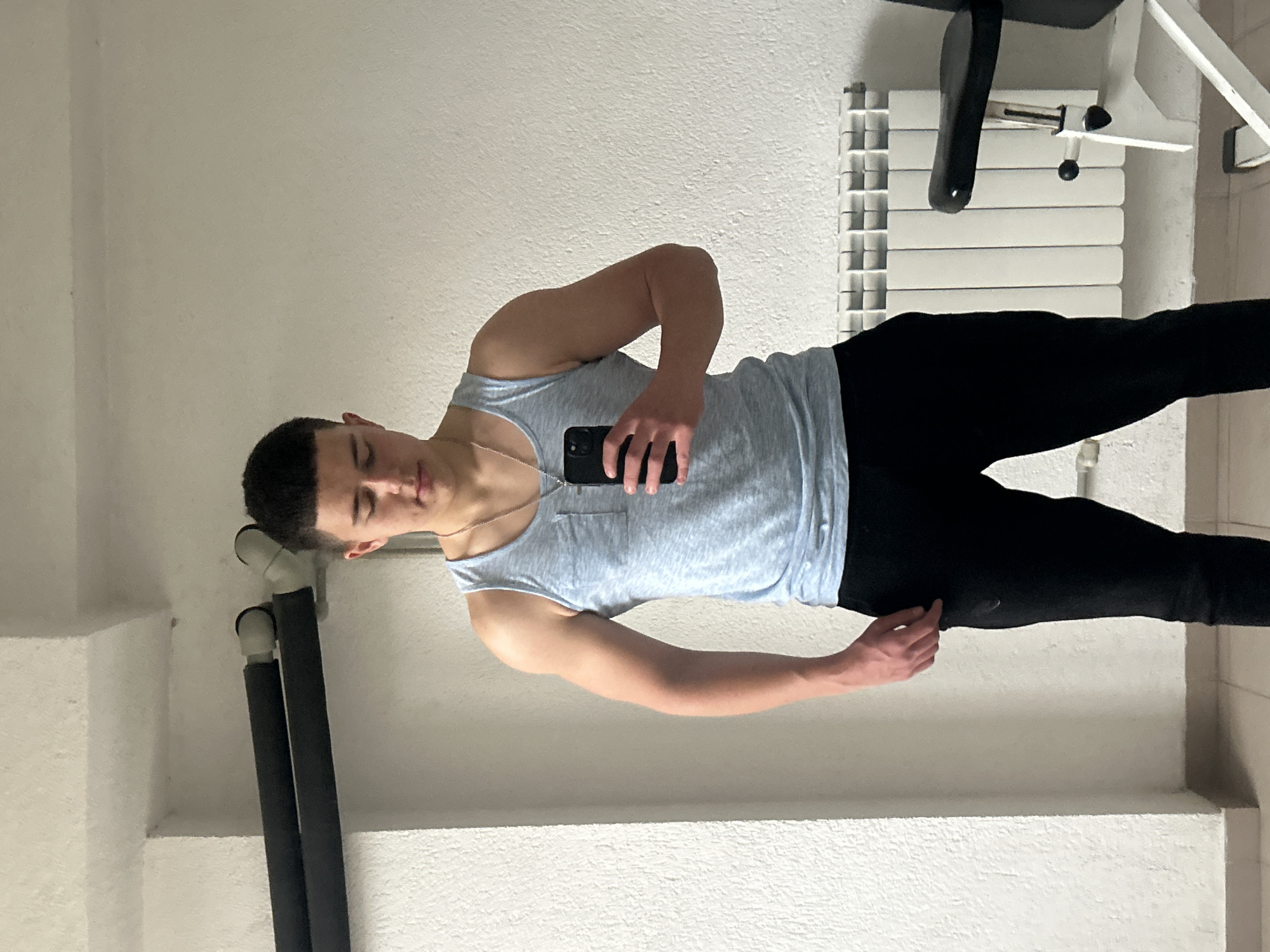A young man, likely in his late teens or early twenties, is depicted in a selfie taken indoors in Zlatograd, Bulgaria. He is positioned centrally, holding a black smartphone in his right hand to capture the image. He has short dark hair and wears a light blue or grey tank top with a small chest pocket and black pants. A thin silver chain or necklace is visible around his neck. His expression is casual, with his eyes slightly closed or squinting as he looks towards the phone.
The background suggests a functional indoor setting, possibly a home gym or a utility room. To the man's right, a white, ribbed radiator or heating unit is prominent, accompanied by elements of gym equipment, including a black padded section and white metal frames. To his left, two dark, elongated cylindrical objects, potentially insulated pipes or equipment parts, are visible against the wall. The walls themselves are a uniform light off-white color with a textured finish. The lighting appears even, typical of an indoor environment. There is no visible text within the image.