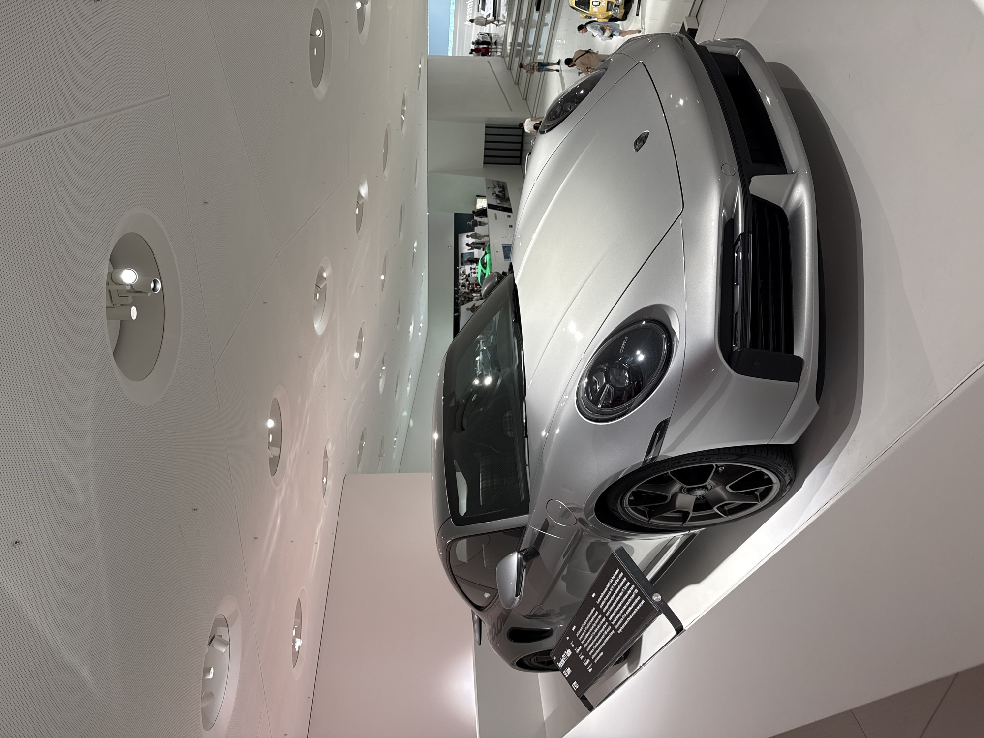The image shows a silver Porsche 911 Turbo S on display in the Porsche Museum in Stuttgart, Germany. The car is positioned on a white pedestal in a well-lit exhibition hall with a modern, minimalist design. The ceiling features a grid of recessed circular lights and ventilation grilles. A black informational plaque is placed in front of the car, providing details about its model and specifications. In the background, several other vehicles are visible, along with displays and architectural elements of the museum. A few museum visitors are also present, looking at the exhibits.