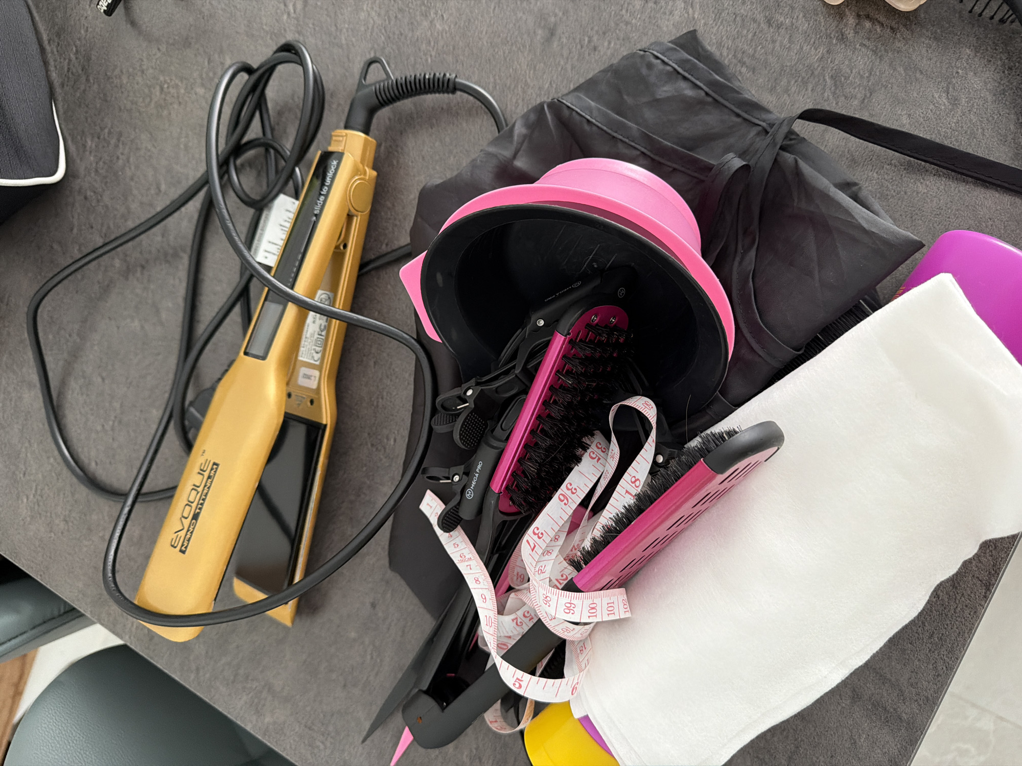 The image shows a collection of hair styling tools laid out on a dark gray, textured surface, likely a table or countertop. The items are clustered together, with some partially overlapping.
Prominently featured is a gold-colored hair straightener with the brand name "EVOQUE NANO TITANIUM" visible. The straightener is plugged in, and its black cord is coiled nearby.
Also present is a pink and black hair brush, a black hair dyeing bowl with a pink rim, and several black hair clips. A white tape measure, marked with red numbers from 90 to 102, is draped across the hair brush and clips. A folded white cloth or towel is situated in the lower left corner. A black fabric garment, possibly a cape or bag, is underneath the dyeing bowl and hair brush. A bright pink object, possibly another hair styling tool, is partially visible in the lower right foreground.
The lighting suggests an indoor setting. There are no people visible in the image.
The presence of hair styling tools does not provide any specific information about the location in Finestrat, Spain.