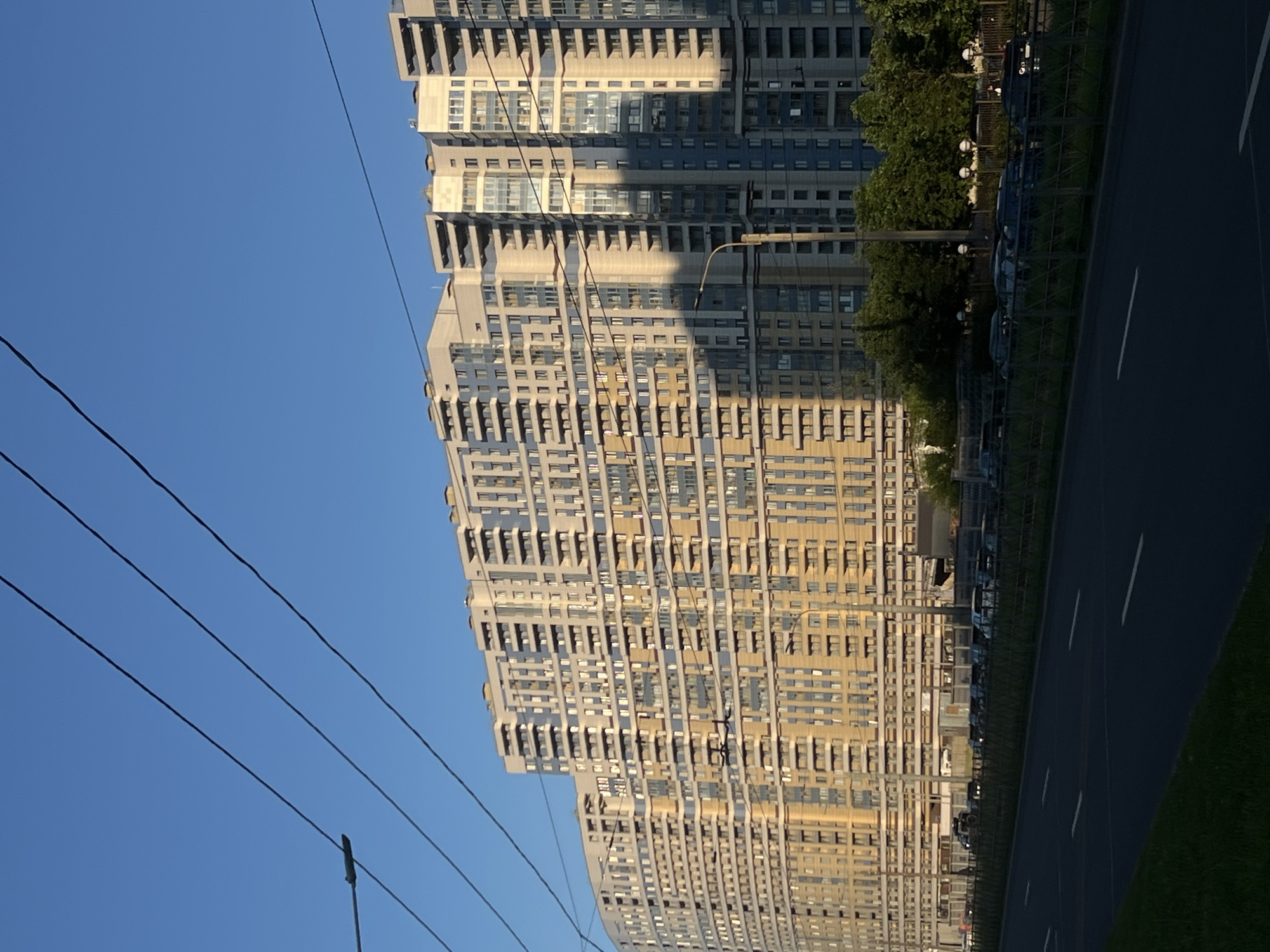 The image captures a broad daytime urban landscape in Saint Petersburg, Russia, specifically along Bogatyrsky Prospekt, featuring the prominent residential complex at addresses such as 2к5. A massive, multi-story apartment building, characterized by its elongated, gently curved facade, dominates the scene. The building's exterior is a blend of light beige/cream and blue-grey panels, with numerous uniform balconies arrayed across its many floors. Sunlight strongly illuminates the building's left and central sections, particularly the lighter-colored surfaces, creating visible shadows on some blue-grey portions and along architectural recesses.
In the foreground, a wide, dark asphalt road, indicative of a major avenue, stretches across the lower third of the frame, showing white lane markings. A low, green-colored metal fence runs parallel to the road on the right, separating it from a section containing parked cars and a row of dense green trees. A tall, dark streetlamp pole stands on the right side of the road, extending upwards towards the building. Overlapping the sky and the upper part of the building are multiple dark, thin electrical wires, consistent with public transport infrastructure (trolleybus lines). The sky above is clear and uniformly blue, devoid of clouds. No individuals are visibly present, but the urban infrastructure and parked vehicles suggest a populated area.
