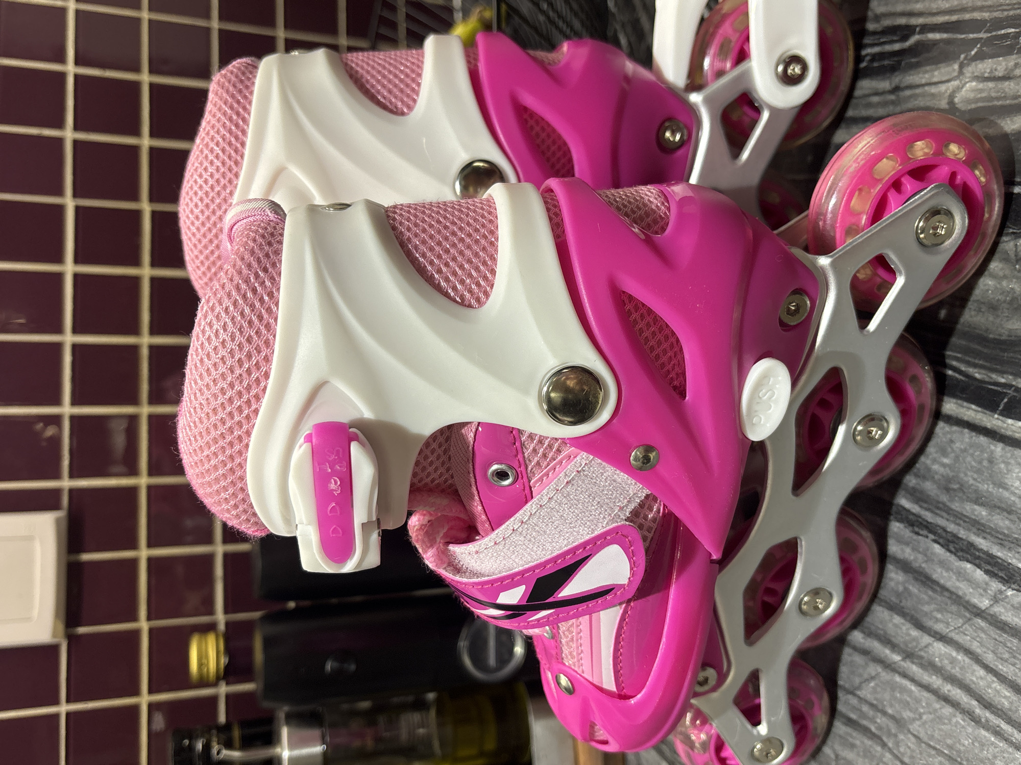 The image shows a close-up, eye-level view of a pair of pink and white adjustable inline skates, likely children's, positioned side-by-side on a grey, textured surface. The skates feature pink mesh fabric uppers, white plastic frames, and clear pink wheels with visible bearings. A pink buckle strap with a white mechanism is visible on the side of the right skate. The background consists of a tiled wall with a dark maroon color and a grid pattern, and out-of-focus kitchen items like bottles are visible on a shelf. The lighting suggests an indoor setting, possibly a kitchen counter. There are no people or actions depicted. The scene is not identifiable as being in Dubai, United Arab Emirates, or any specific city.