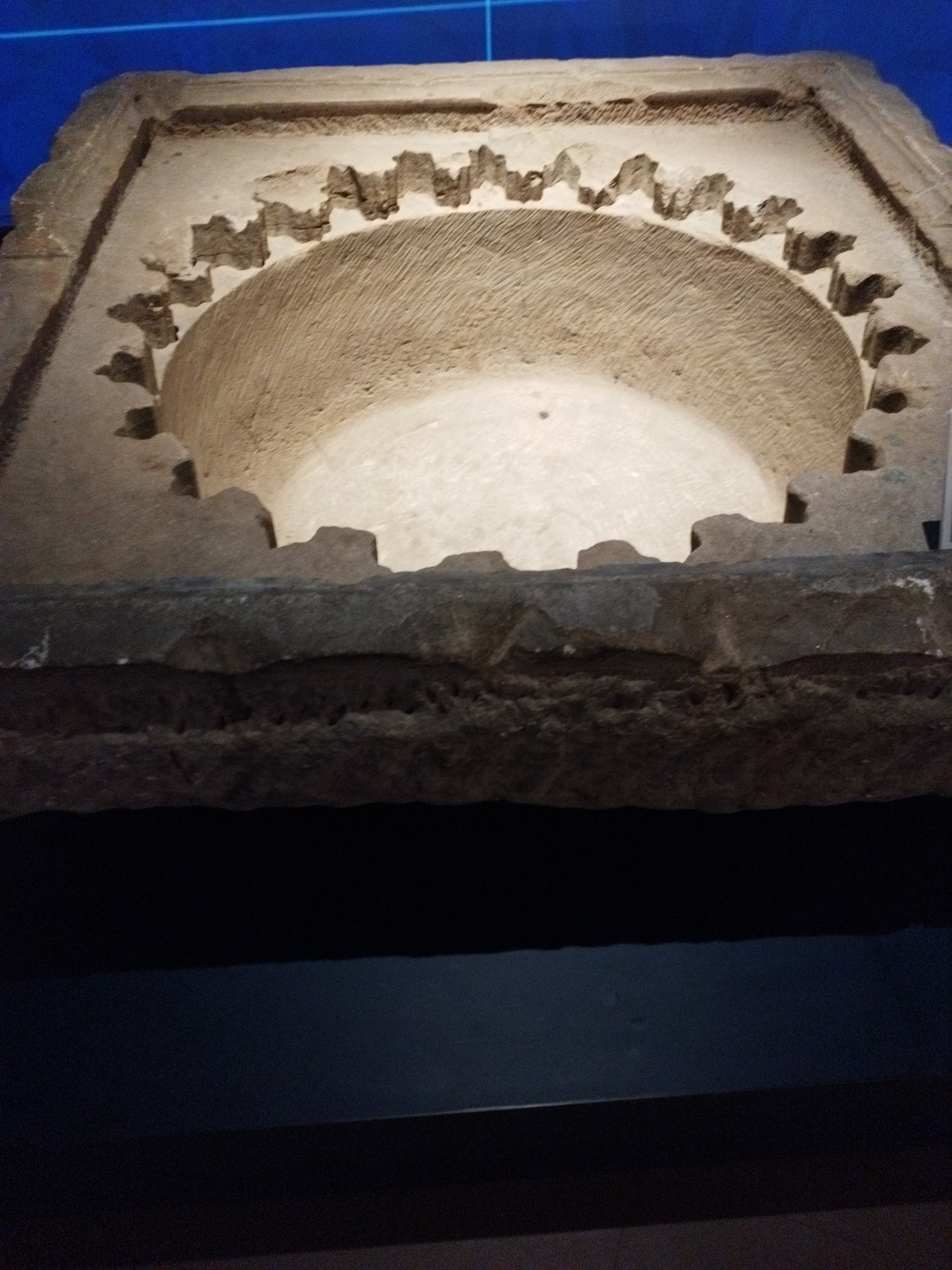 The image shows a close-up, angled view of an ancient stone artifact, possibly part of an architectural element, set against a dark background. The artifact is rectangular with a carved, semicircular niche in the upper portion. The edges of the niche are decorated with a series of triangular or pointed protrusions, resembling teeth or crenellations. The surface of the stone within the niche is rough and exhibits parallel striations, indicative of tool marks. Below the niche, the stone is flat and appears to be a heavy slab, also showing signs of wear and damage. The overall impression is of an aged and weathered piece of stone carving, presented as an exhibit. The background is dark, with a hint of a blue surface at the top, suggesting it might be displayed in a museum or gallery setting.