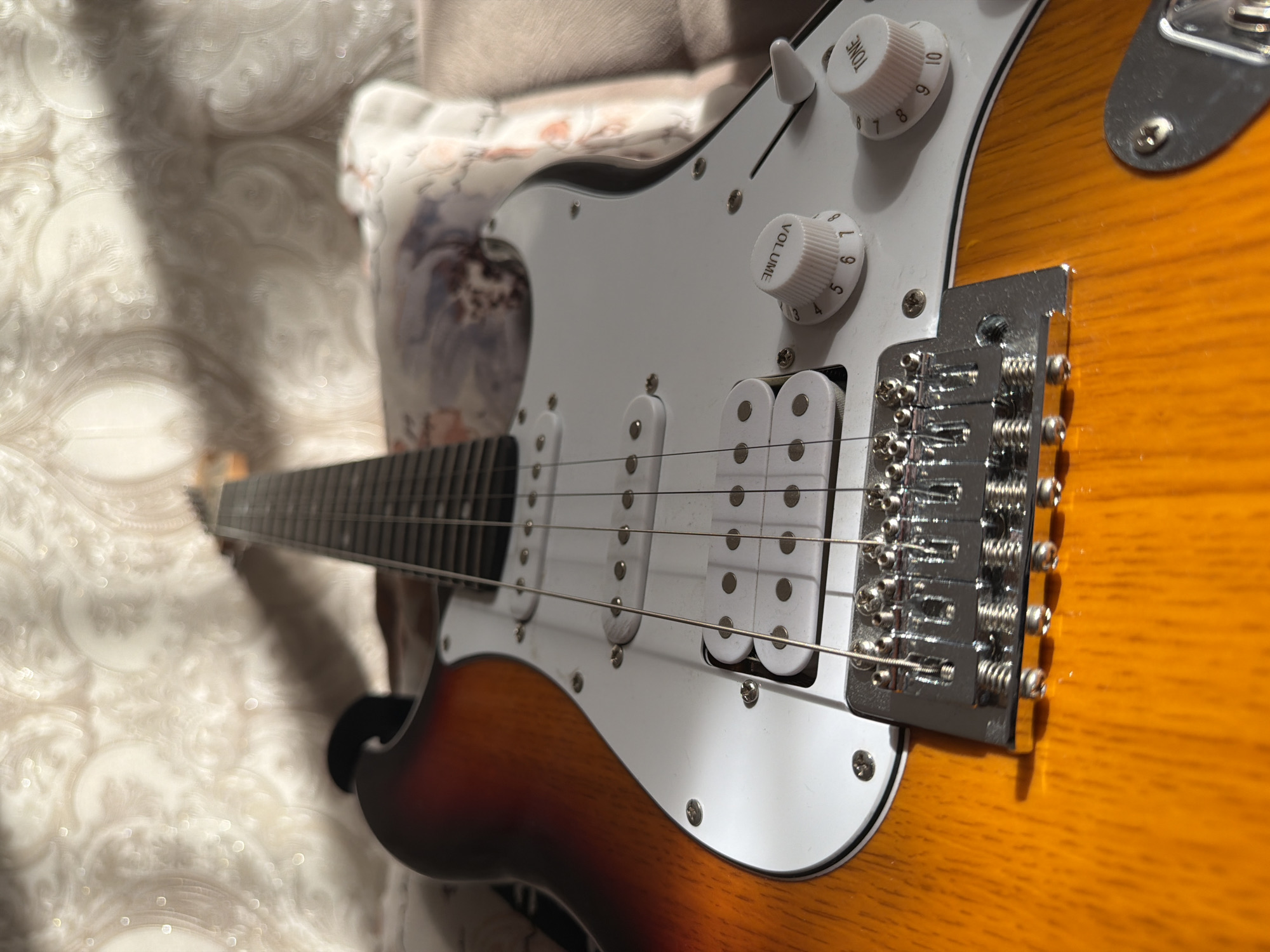 A close-up, low-angle shot captures an electric guitar lying on its back. The guitar has a sunburst finish, transitioning from a deep red at the top to a bright orange at the bottom. Its white pickguard is visible, featuring two white pickups and three white knobs, two of which are labeled "VOLUME" and "TONE" with numbered markings. The metal bridge and strings are prominent in the foreground, reflecting light. The background shows a patterned wallpaper with light and shadow play, suggesting an indoor setting, possibly a home. The specific location in Baku City, Azerbaijan, cannot be determined from the visual information provided.