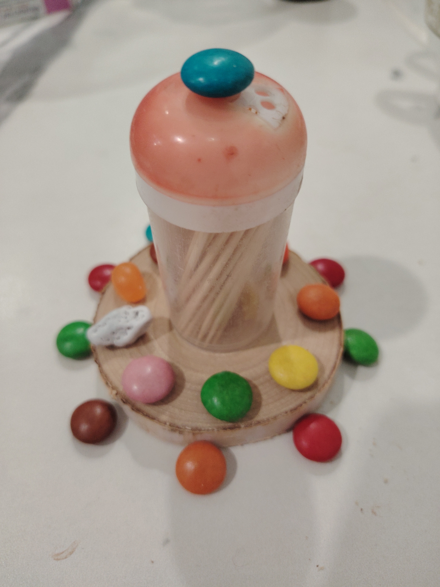 A close-up shot captures a toothpick holder crafted to resemble an ice cream cone. The "cone" is a clear plastic container filled with wooden toothpicks. It sits atop a small, circular wooden base. Surrounding the base are scattered multi-colored candies. The scene is situated on a light-colored surface. There is no verifiable information to support a location in the city of Sarapul, Russia.