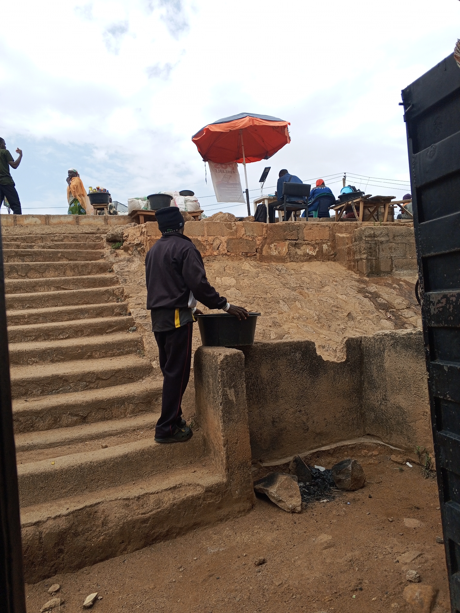 Gwong, Nigeria. A man in a black tracksuit and hat ascends stone steps with a black bucket. Behind him, a raised area houses several individuals; one works at a table under an orange umbrella. At the bottom of the steps is a small area with charred rocks and debris. A partial gate is visible on the right side of the frame.