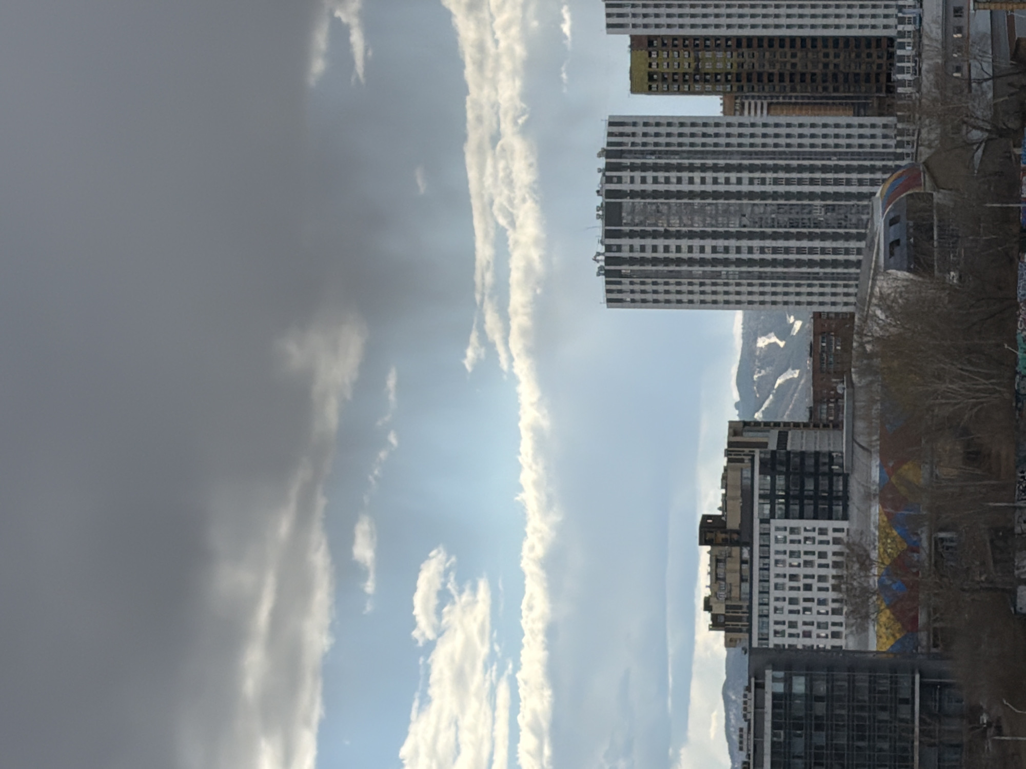 This image captures a cityscape in Krasnoyarsk, Russia, under a dramatic, partially cloudy sky. The scene is dominated by several tall, modern residential buildings, many with numerous windows, some appearing to be under construction with visible yellow insulation panels. Behind these urban structures, snow-capped mountains are visible in the distance, contrasting with the built environment.
The sky is a prominent feature, transitioning from a dense, dark grey cloud mass on the left to brighter, scattered white clouds and patches of clear blue sky on the right. Beams of sunlight are visible breaking through the clouds, creating crepuscular rays. Bare trees, indicative of autumn, winter, or early spring, are visible in the lower right foreground, with a colorful mural featuring geometric patterns in red, yellow, and blue on a building wall behind them.
The photograph depicts a daytime scene, likely late morning or afternoon, given the bright sun. No people or specific activities are discernable. There is no visible text in the image. The overall impression is a blend of urban expansion and natural landscape, characteristic of a city nestled near mountains.