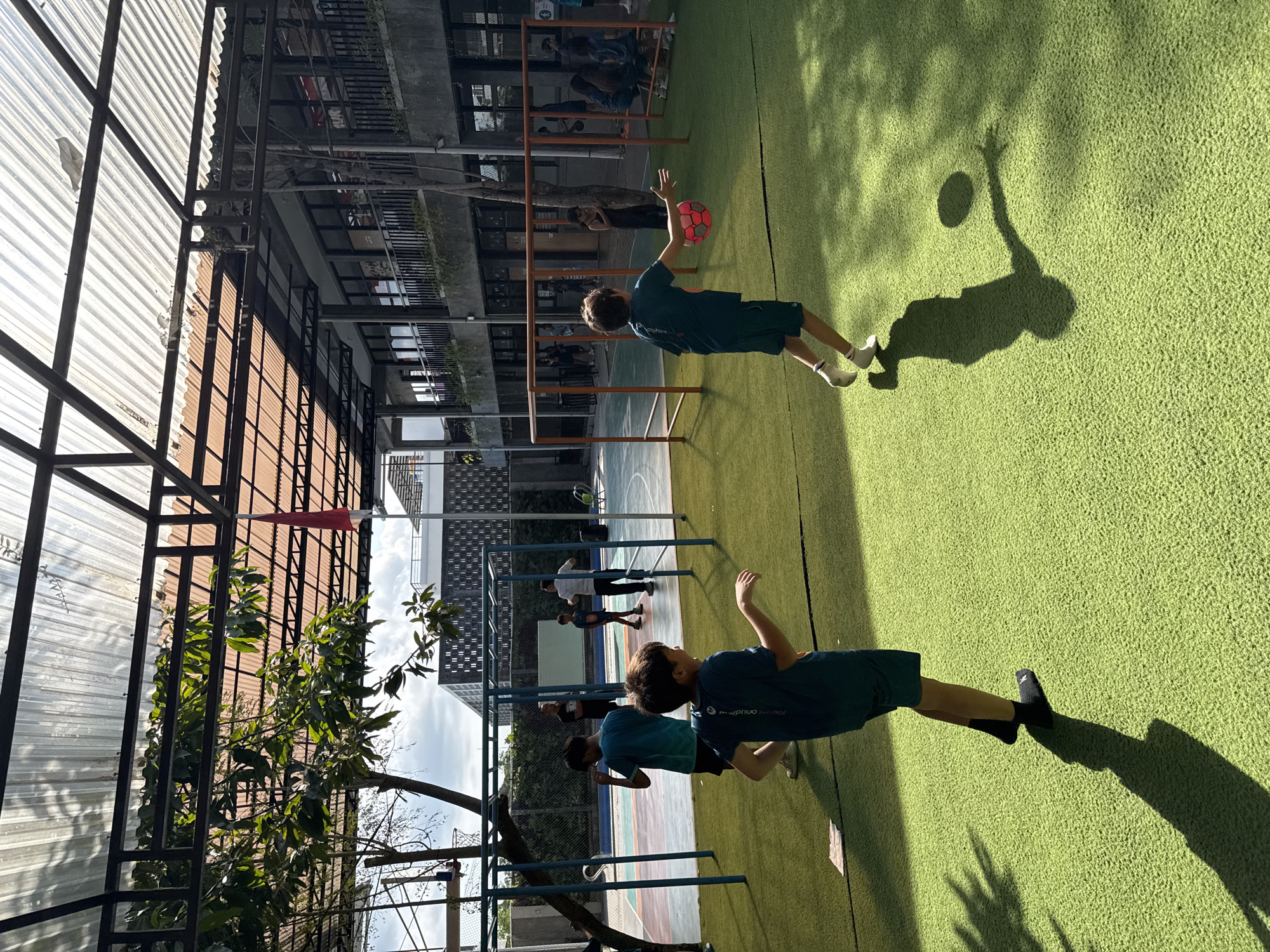 The image depicts an outdoor sports field with artificial turf in North Kuta, Indonesia. Several children are present on the field, engaged in activities. In the foreground, a boy in a dark teal shirt and shorts is running with a red and white patterned ball in his left hand, his body angled as if throwing or preparing to throw it. His shadow is cast on the grass to his right. Behind him, another boy, also in a dark teal shirt and dark shorts, stands with his left arm bent and his right arm extended slightly, looking towards the left of the frame. Further back, other children are visible, some near a set of blue monkey bars and others near an orange soccer goal. In the background, a multi-story building with balconies and windows is visible, and beyond that, a cloudy sky. The field is covered by a translucent corrugated roof supported by a metal frame, casting dappled shadows on the grass. A red and white flag is mounted on a pole to the left of the building.