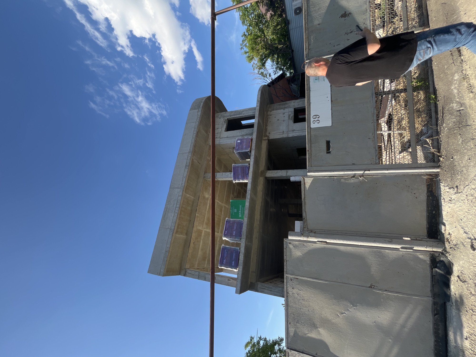 The image captures a sunlit construction site, featuring a concrete building in the early stages of construction, a man, and a clear blue sky.
The central subject is a multi-story concrete building with an distinctive, curved roofline. Several stacks of construction materials, wrapped in purple and one in green plastic, are visible on one of the building's upper levels. The green package clearly displays the Cyrillic text "ВКБлок". The purple packages feature a logo that appears to be "СК" or "CK". A brown metal pole, possibly scaffolding or support, extends vertically in front of the building.
On the far right, a man is partially visible, seen from the waist up. He is wearing a black t-shirt and blue jeans, standing next to a concrete wall and a metal gate. He appears to be looking towards the left side of the frame. On a white board attached to the concrete wall, the number "39" is clearly visible.
The scene is set outdoors on a bright, sunny day, indicated by the vibrant blue sky with scattered white clouds and the sharp shadows cast. Lush green trees are visible in the background and peeking into the foreground, suggesting a natural environment surrounding the construction. The presence of Cyrillic text on the packaging aligns with the provided location context of Krasnodar, Russia. The overall atmosphere is that of an active construction zone under clear weather conditions.