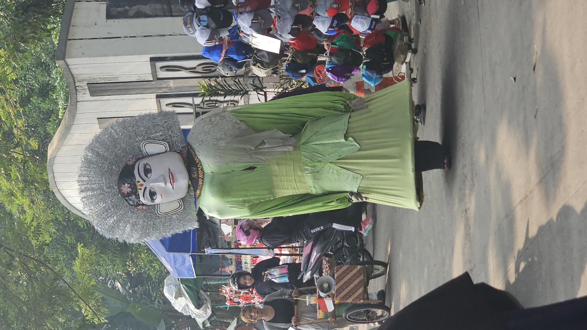 The image captures a street scene in Depok, Indonesia, with a large, elaborately dressed Ondel-ondel puppet as the central focus. The puppet, a traditional Betawi folk art figure, is predominantly dressed in a vibrant lime green costume, complete with a wide sash and flowing skirt. Its head is painted white with a striking resemblance to a human face, adorned with black hair and a large, silver, feathery headdress. The puppet is standing on a paved street, with its lower half obscured by its voluminous skirt.
Several people are visible in the background, engaged in various activities. To the left, a woman with blonde hair is pushing a cart equipped with a megaphone and a small Indonesian flag. Behind her, two young women wearing helmets are on a motorcycle. Further in the background, stalls display an array of colorful hats. The environment suggests a lively outdoor market or street festival, with tents and structures indicating commercial activity. The lighting indicates it is daytime, with sunlight casting shadows on the ground.