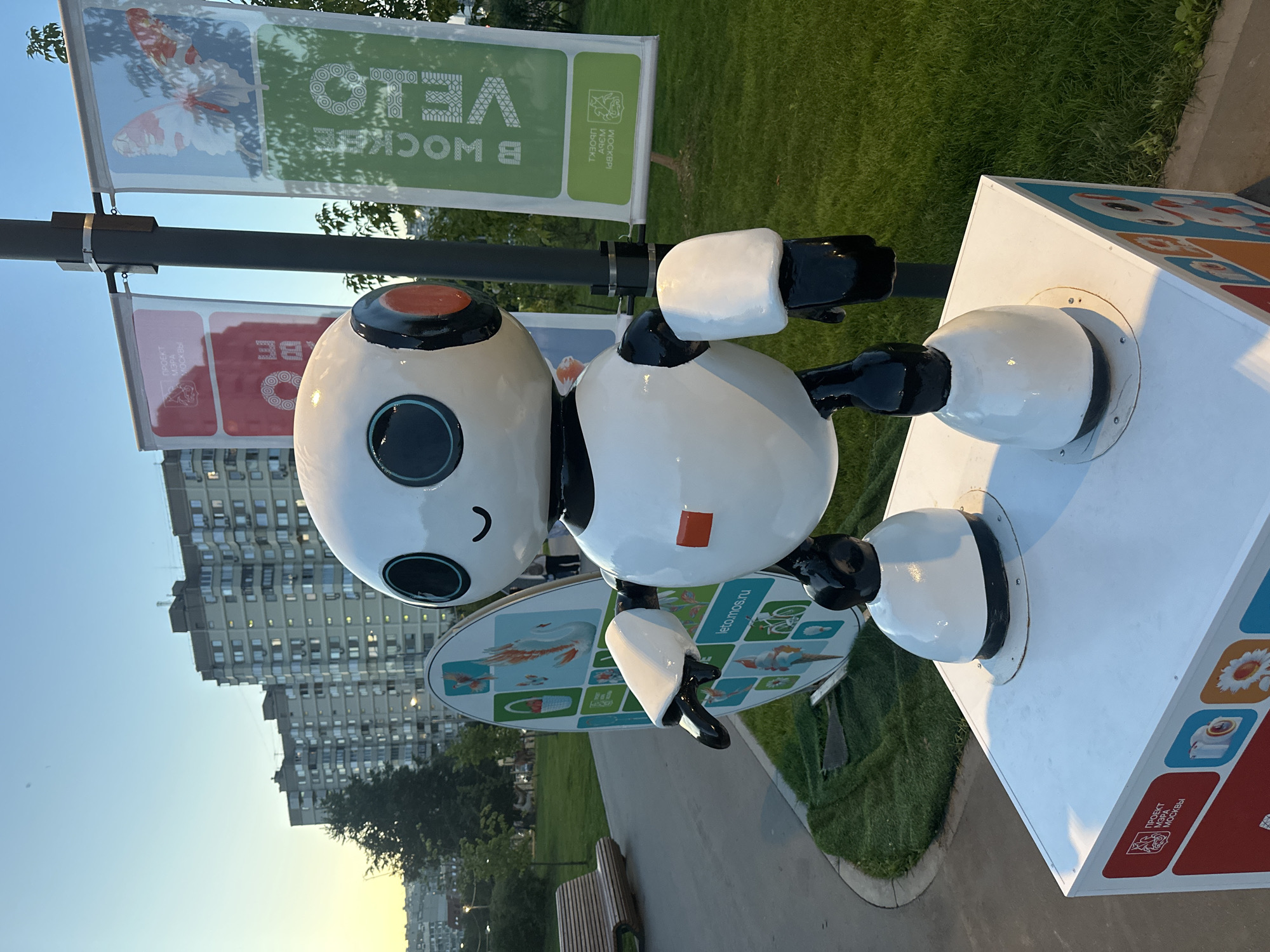 A white and black robot stands on a white pedestal in a park, with tall residential buildings in the background. The robot has a round head with large, dark blue eyes and a small smile. It has orange accents on its head and a red square on its chest. The pedestal it stands on is decorated with colorful images and text, including Russian phrases like "ПРОЕКТ МЭРА МОСКВЫ" (Mayor's Project of Moscow).
Behind the robot, a lamppost displays two banners. The left banner is red with text and an emblem, while the right banner is green and features an image of a butterfly and text. A large circular display with various colorful illustrations, including a swan and ice cream, is positioned to the left of the robot.
The park environment includes a paved walkway, lush green grass, and a wooden bench. Trees are visible in the mid-ground. The sky is a clear blue, transitioning to a warm, soft yellow hue near the horizon, suggesting it is either early morning or late evening. The overall scene appears to be an outdoor public space in Moscow, Russia, possibly a park or recreational area designed for community engagement, indicated by the "Mayor's Project of Moscow" branding. There are no people clearly visible in the immediate vicinity of the robot, though figures can be seen in the background.