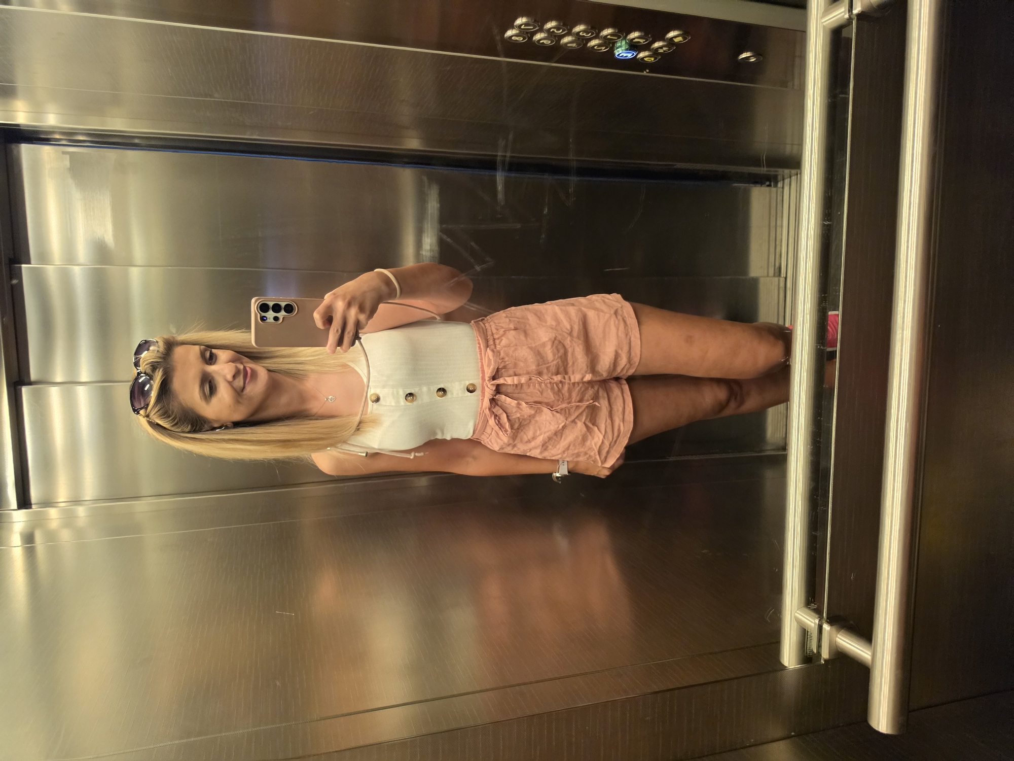 A blonde woman with sunglasses perched on her head is taking a mirror selfie in an elevator. She is wearing a white sleeveless top with buttons and pink shorts tied with a ribbon. The elevator walls are made of stainless steel, and there is a control panel with buttons on the right side. In the foreground, a metal handrail is visible. The overall impression is of a casual moment captured in a mundane urban environment.
