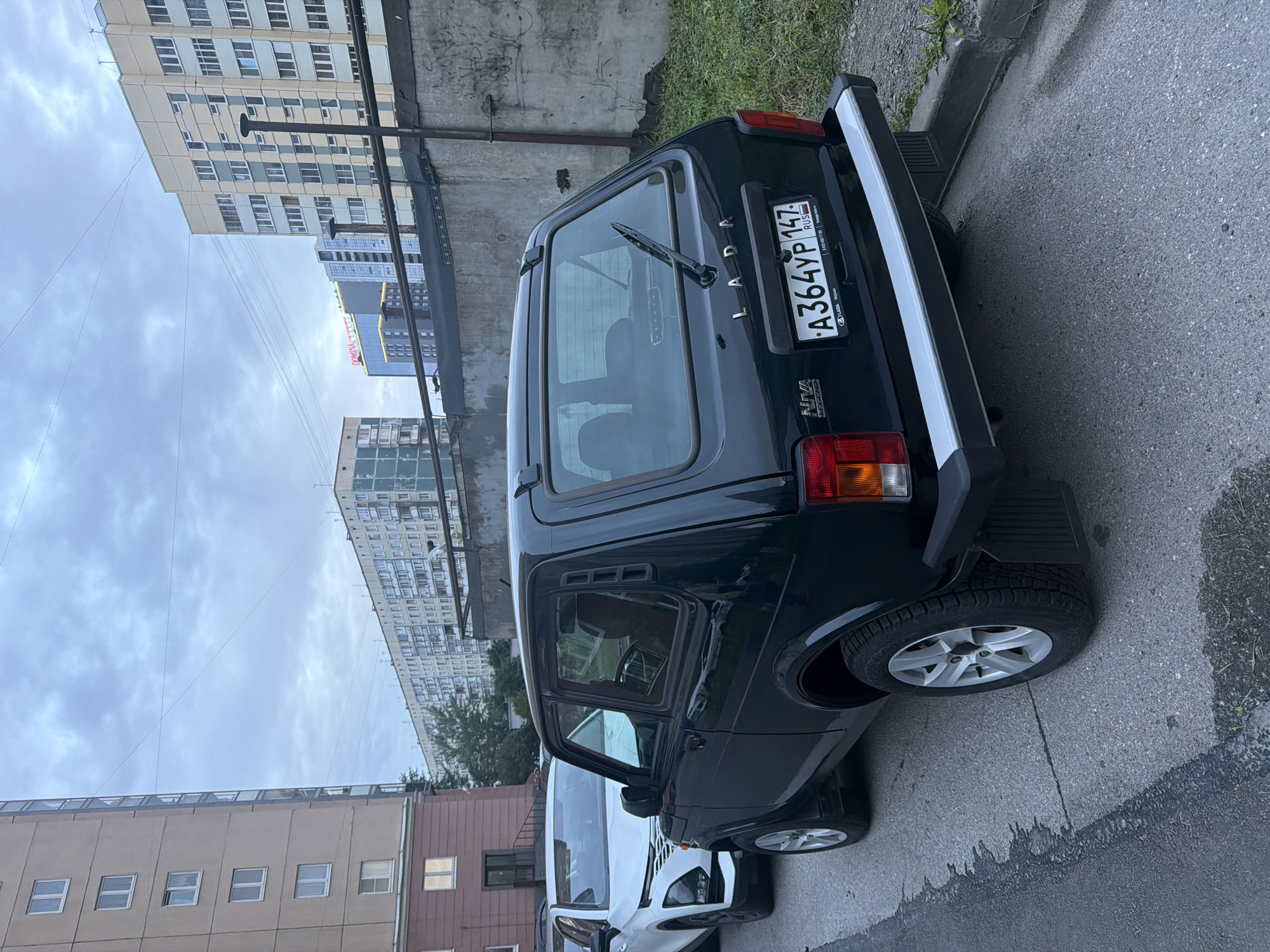 A dark green Lada Niva Legend SUV is parked on an asphalt street next to a white SUV. The Lada's license plate reads "A364YP 147 RUS". In the background, there are several multi-story apartment buildings under a cloudy sky. A concrete wall runs along the right side of the image, covered in graffiti.