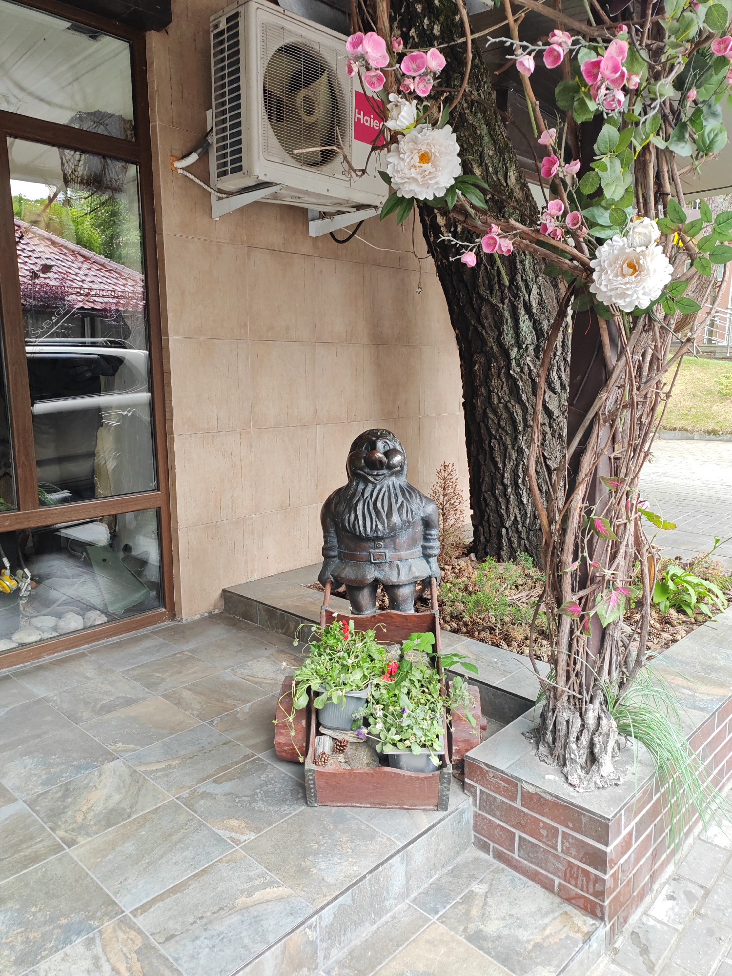 The image depicts an outdoor scene in Svetlogorsk, Russia, likely an entrance to a building. In the foreground, a tiled patio is visible. To the left, a large window with a wooden frame reveals a reflection of trees and a roof with red tiles. An air conditioning unit is mounted on a beige tiled wall above the window.
A bronze garden gnome statue stands on the patio, holding a wooden cart filled with potted plants, including some with red flowers and green leaves. The gnome is detailed with a beard, tunic, and belt.
Behind the gnome and to the right, a tree trunk is prominent, adorned with artificial pink and white flowers and branches that weave around the trunk and a supporting structure made of vertical brown rods. The base of the tree is surrounded by pinecones and some low-growing greenery.
The right side of the image shows a brick planter, with a section of the tiled patio continuing around it. Green grass and paved walkways are visible in the background. The overall impression is of a decorated entrance area with a mix of natural and artificial elements.