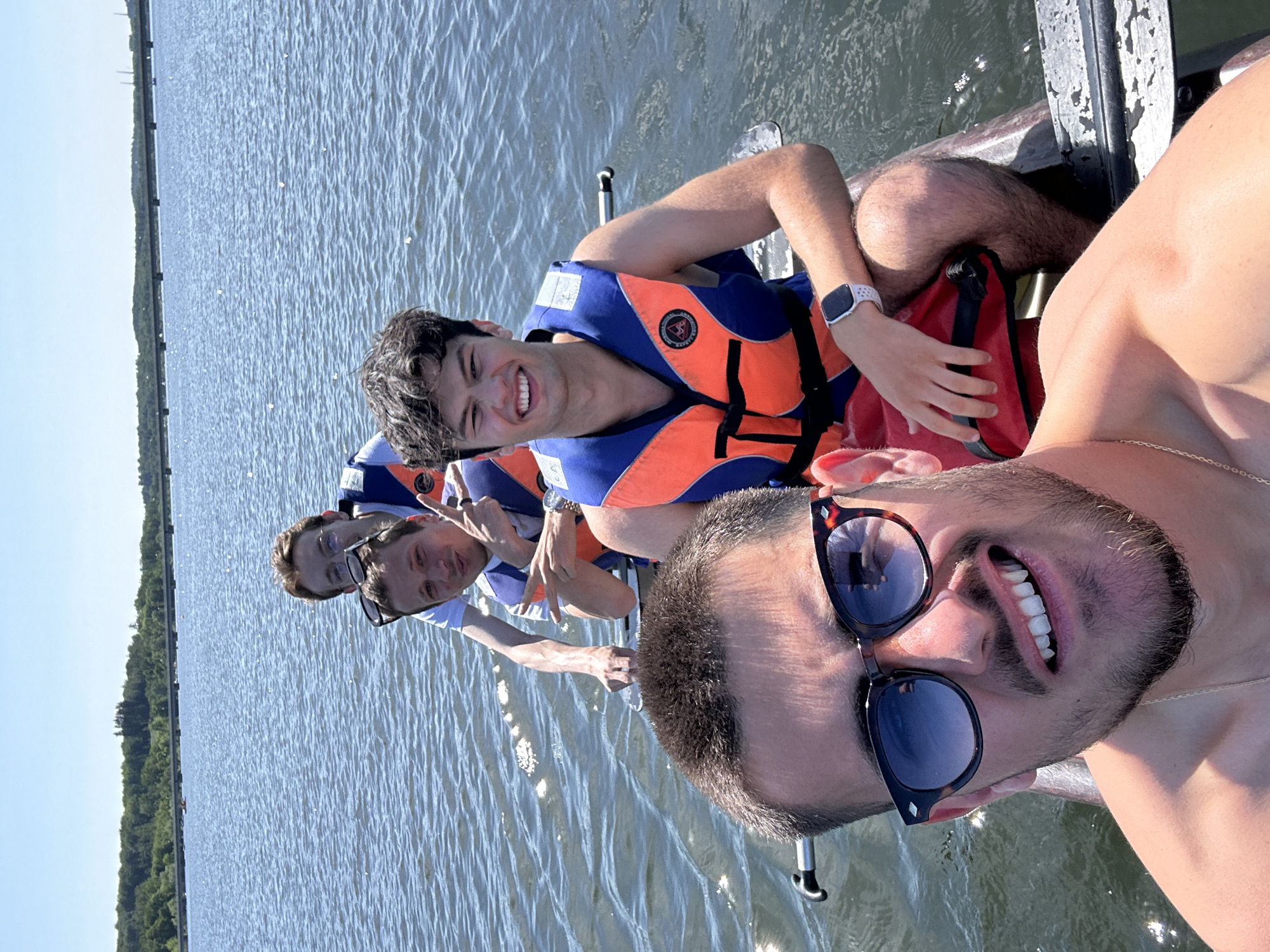 Four people are riding in a boat on water. All are wearing life vests except one man in the foreground. One man in the foreground is wearing sunglasses and a gold chain. Three men in the back are wearing life vests. A red and black bag is in the boat. The background shows a line of trees on land. The location is Möhnesee, Germany.