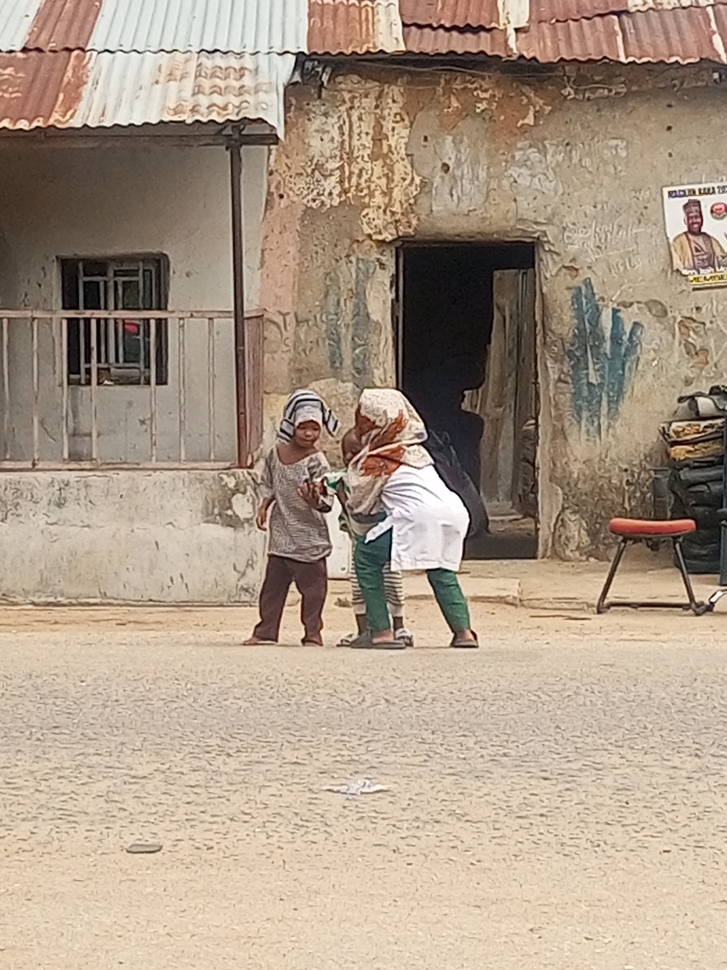 Three children stand on a paved road in Maiburiji, Nigeria, in front of a dilapidated building. One child, wearing a patterned shirt and pants and a striped head covering, faces the camera. Another child, partially obscured, wears a white top, patterned pants, and a headscarf. The third child is largely obscured. A building with a rusty metal roof and peeling paint serves as the backdrop. A dark doorway is visible in the building's facade, next to a poster on the wall. To the right of the doorway, a stool sits near stacks of tires.