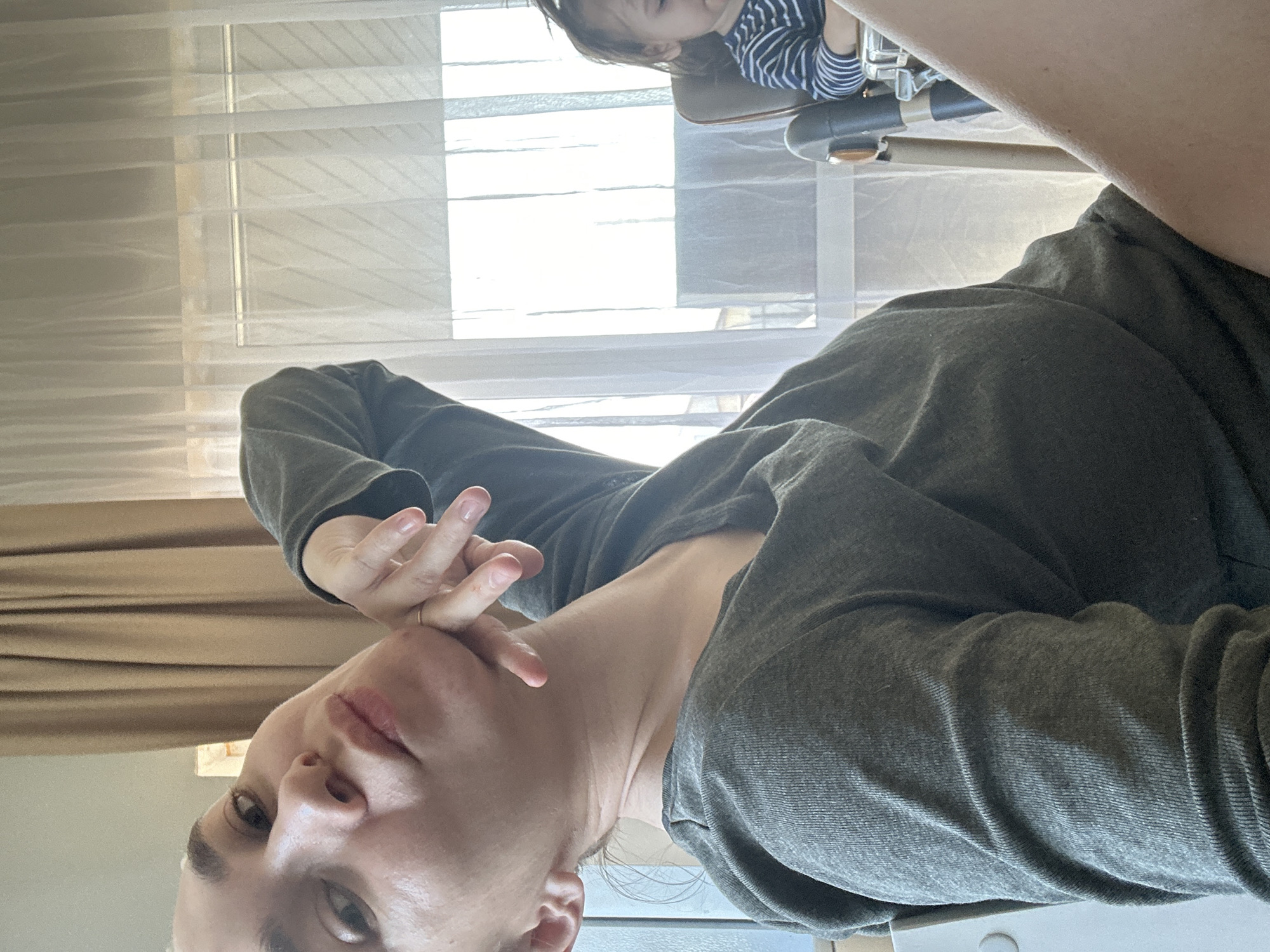 A woman with short, light hair and a gray long-sleeved shirt is facing the camera with a downward tilt. She is touching her face with her hand. A child with dark hair wearing a striped shirt is visible in the background, seated in a high chair. A window with sheer curtains is behind them. The location is unclear, and there is no information to conclude that the location is in the city of Kharnavy, Russia.