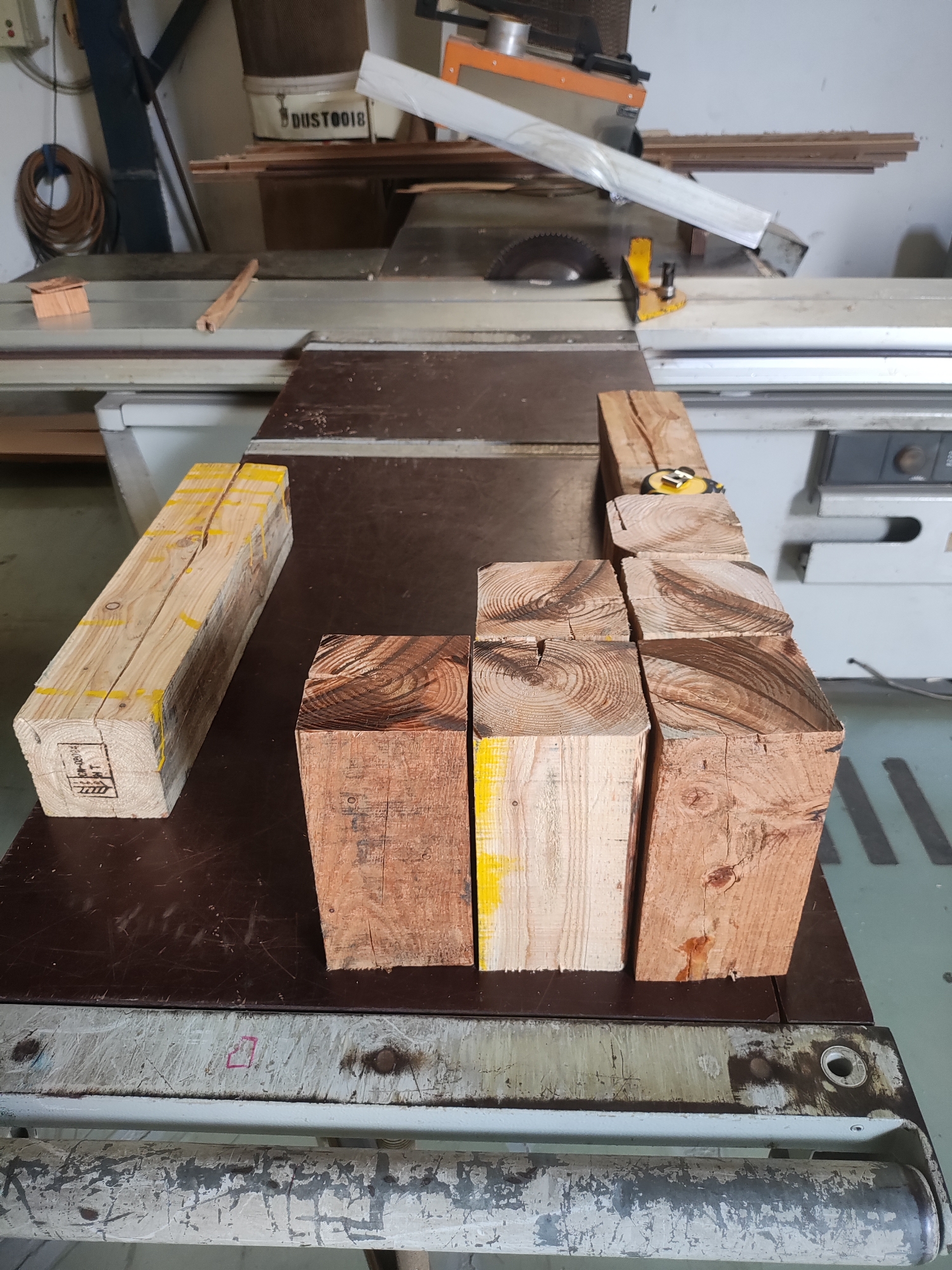 The image shows a workshop with woodworking equipment and materials. In the foreground, several pieces of lumber are arranged on a dark brown tabletop. Some of the lumber pieces are rectangular blocks, while one is a longer, square-profiled piece marked with yellow paint. These pieces appear to be raw wood, possibly for crafting or construction.
Behind the tabletop, a large woodworking machine, likely a table saw, is visible. It has a visible saw blade and yellow safety guards. The machine is white with some gray accents. To the left of the table saw, a coiled brown cord is hanging on a wall. Further in the background, stacks of lumber are visible, along with a white paper roll. A dust collector bag labeled "DUST0018" is also present. The walls of the workshop appear to be light-colored.
The scene suggests an active or recently used woodworking area. The presence of lumber, tools, and the woodworking machine indicates a place where woodworking activities are performed. The overall environment appears to be an indoor workshop space. Without further information or distinct landmarks, it is not possible to determine the exact location within East Jakarta, Indonesia, from the visual content alone.