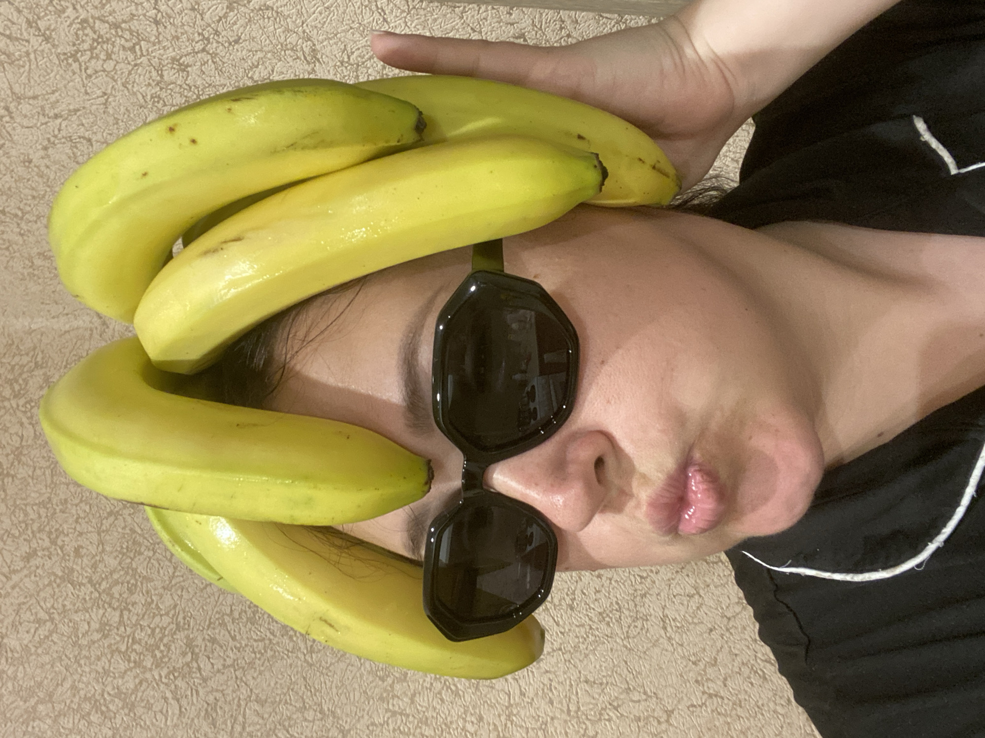 A person is lying on their back, wearing sunglasses and with bananas arranged on their head. The person is wearing a black top with white trim. The background is a beige surface with a slightly mottled pattern. There is no evidence to suggest the location is Sochi, Russia.