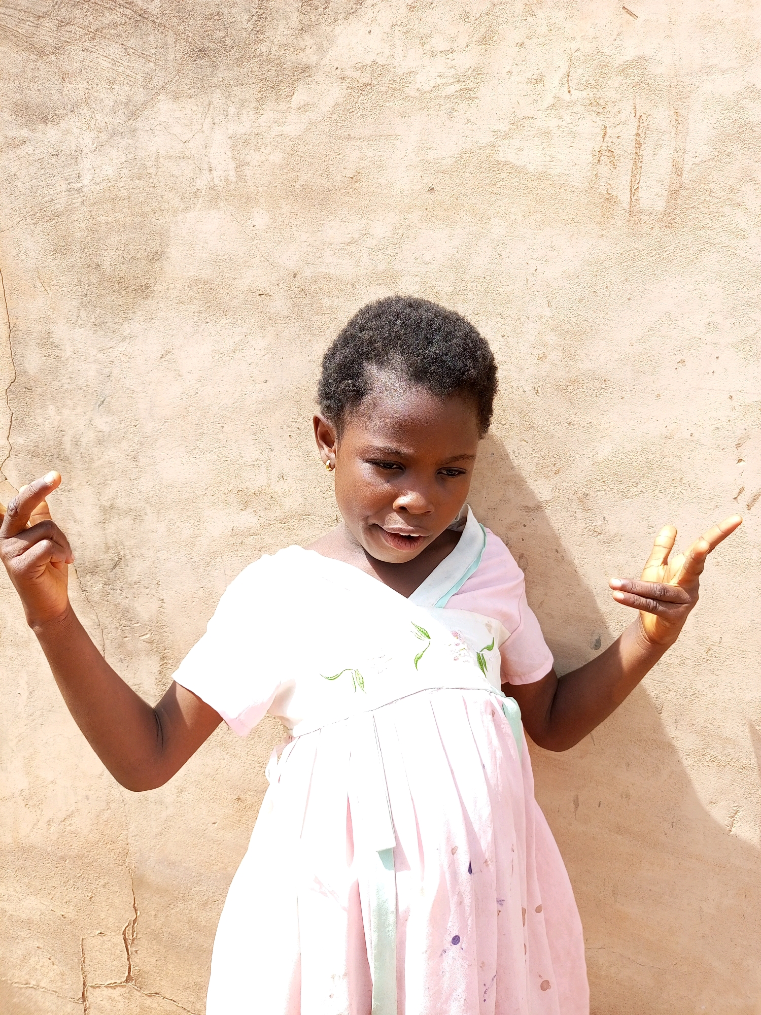 A young Black girl stands in front of a textured, light brown wall. She is wearing a light pink dress with a white collar and subtle green floral embroidery. Her hair is short and dark. Her left arm is bent at the elbow, with her hand raised and fingers curled. Her right arm is also bent, and her hand is held up, with her index and middle fingers extended, and her thumb and ring finger touching. The wall behind her has visible cracks and indentations, suggesting an outdoor or aged structure. Sunlight casts a shadow of the girl onto the wall to her right. The overall scene appears to be in an outdoor setting, possibly a courtyard or street in Tempure, Nigeria, given the texture of the wall and the lighting conditions.