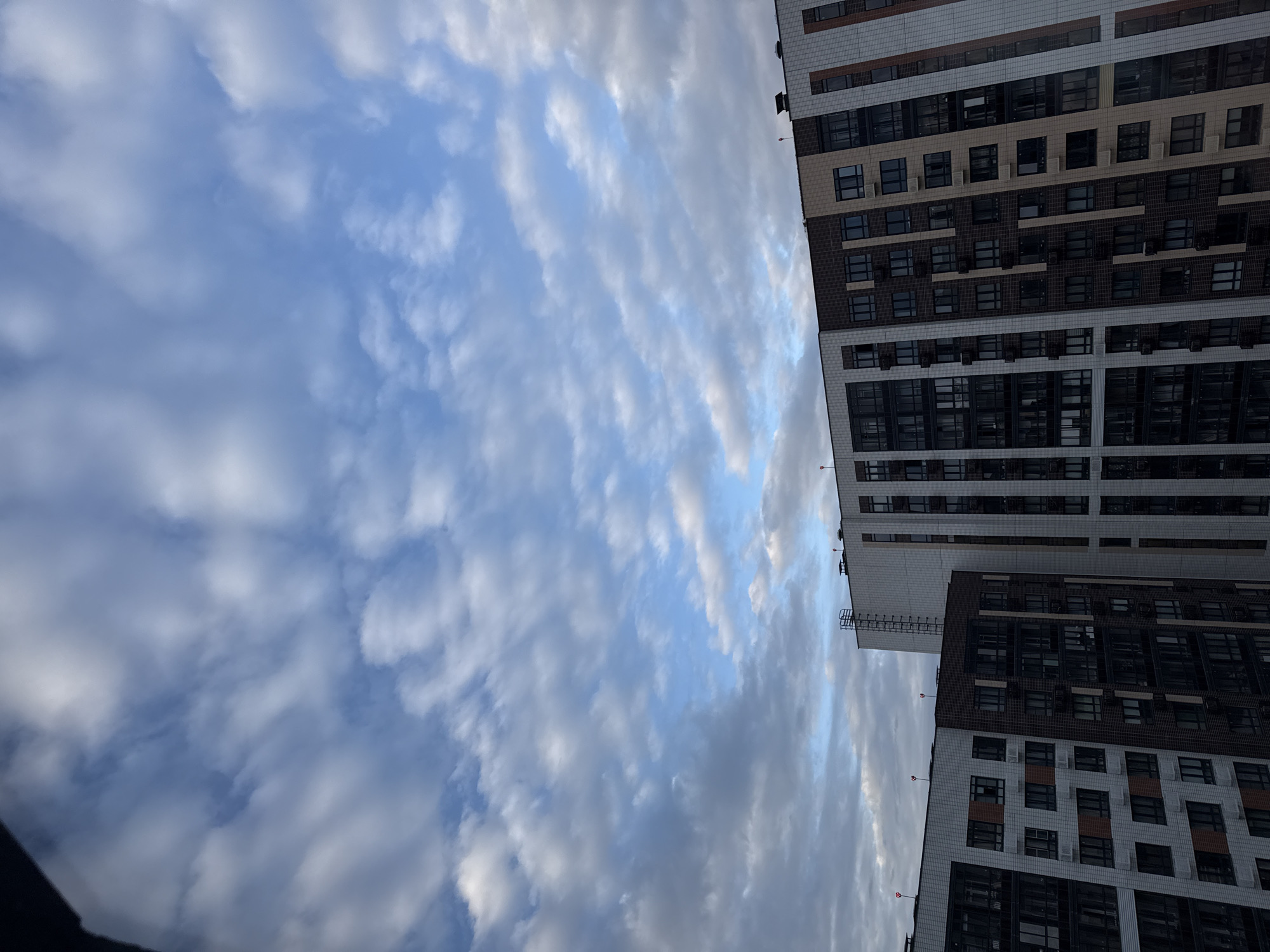 The image shows a view from a window looking up at two modern apartment buildings against a cloudy sky. The buildings are multi-story structures with a facade of white, dark brown, and tan colored tiles, and numerous windows. Some windows are dark, suggesting they are occupied or have dark curtains, while others appear reflective. Air conditioning units are visible outside some windows. The top of the buildings feature some architectural details and what appear to be red lights. A metal ladder is attached to the side of the taller building. The sky is filled with scattered clouds in varying shades of white and gray. The scene is likely captured in the city of Lyubertsy, Russia, given the architectural style of the buildings, which are characteristic of newer residential developments in the area. There are no people visible in the frame.