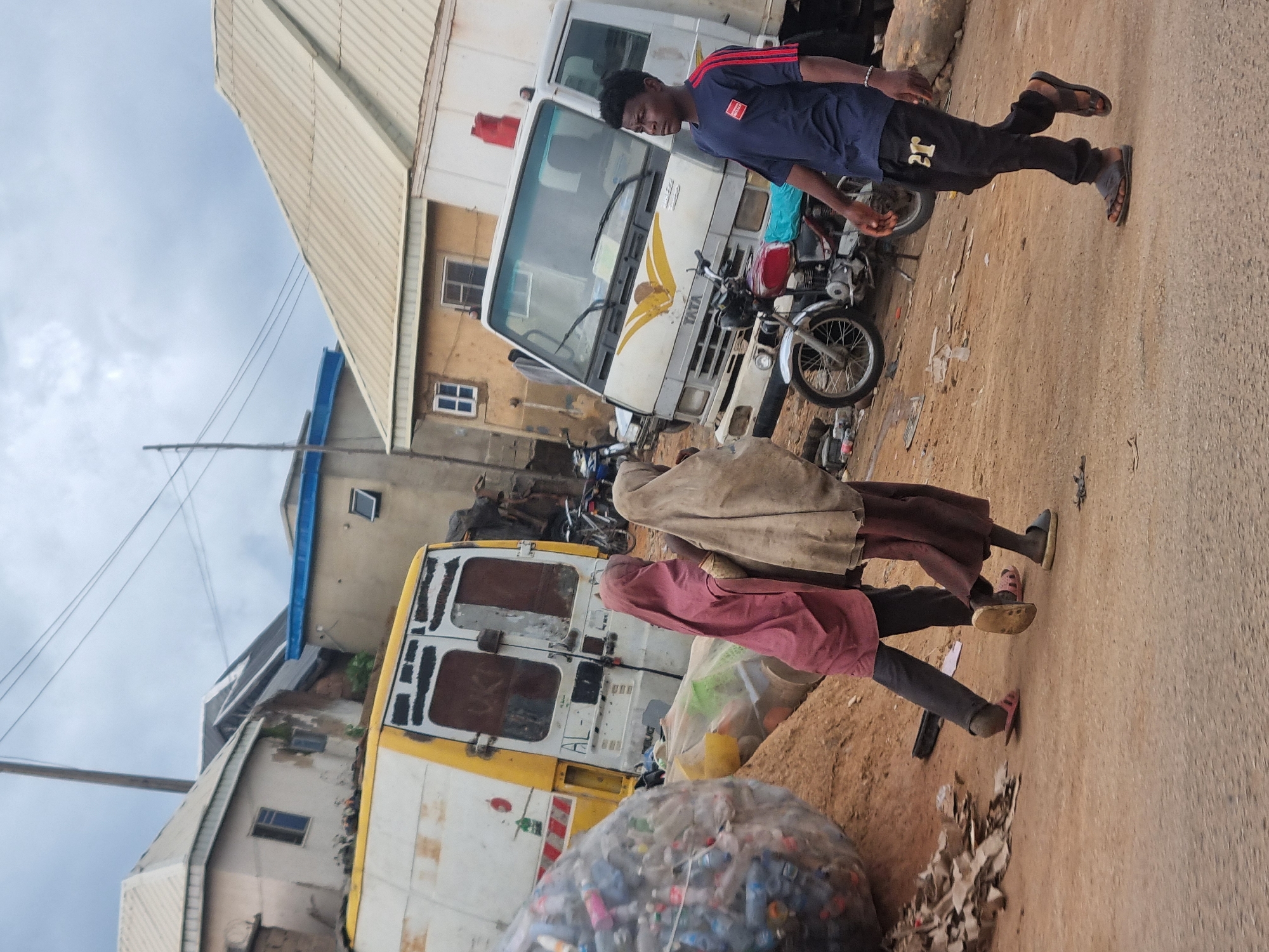 In the city of Gwong, Nigeria, a dusty street is lined with buildings, some with corrugated metal roofs. Two children, one wearing a maroon hijab and the other a beige hijab, walk away from the camera, their faces obscured. To their left, a large, clear plastic bag is filled with plastic bottles. Behind the children, a white van with yellow trim is parked, and further behind it, a white truck with a yellow emblem is visible. A red motorcycle is parked next to the truck. A young man in a blue shirt and black pants walks past the truck and motorcycle, also walking away from the camera. The sky is overcast.