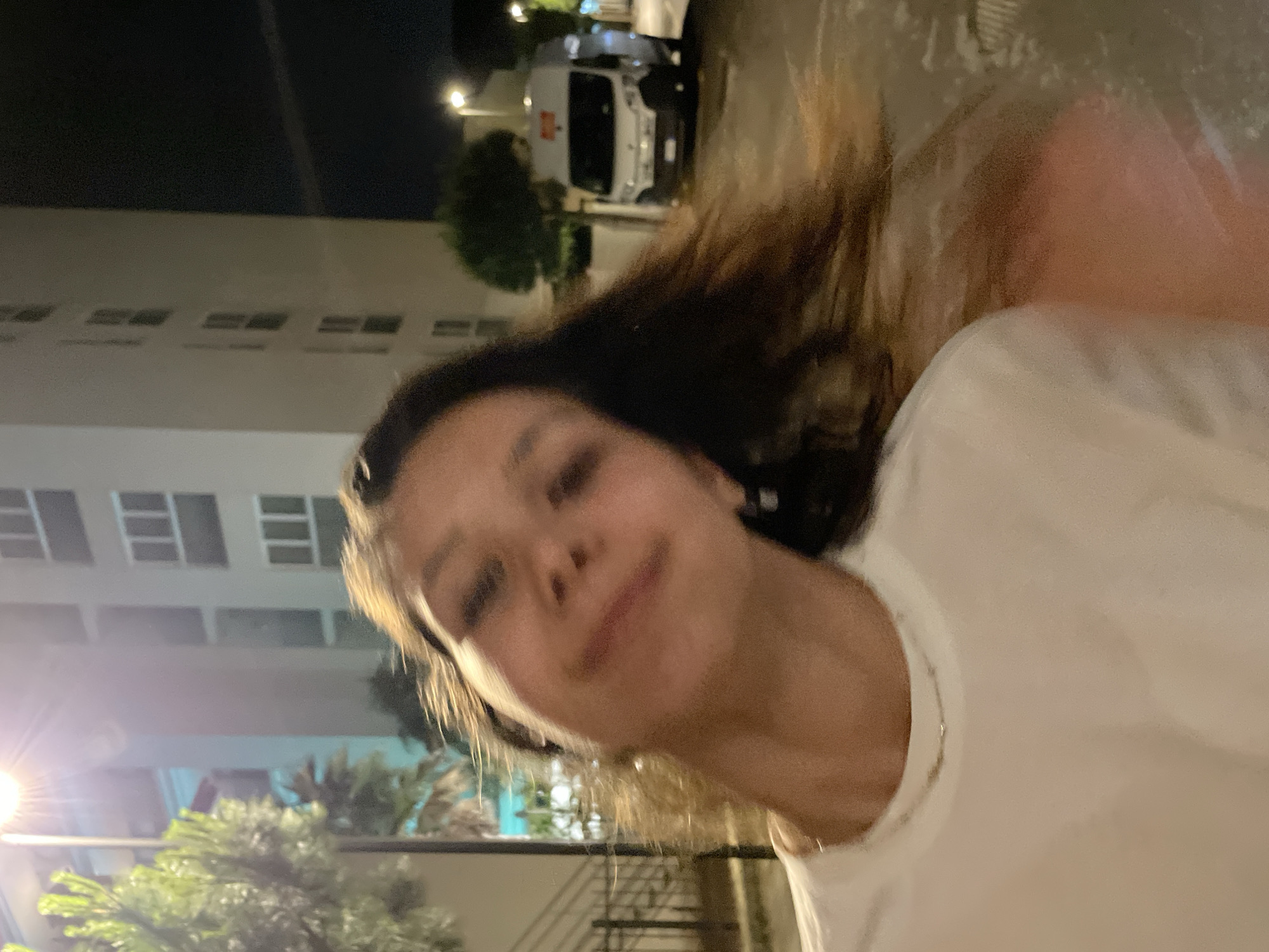 A photograph taken at night appears to be a selfie of a person with tan skin and long brown hair wearing a white shirt and a gold necklace. Behind the person is an out-of-focus building with multiple floors and windows lit by street lights. Trees are partially visible in the foreground, and a white truck is parked near the building. The location is identified as the city of Jesolo, Italy.