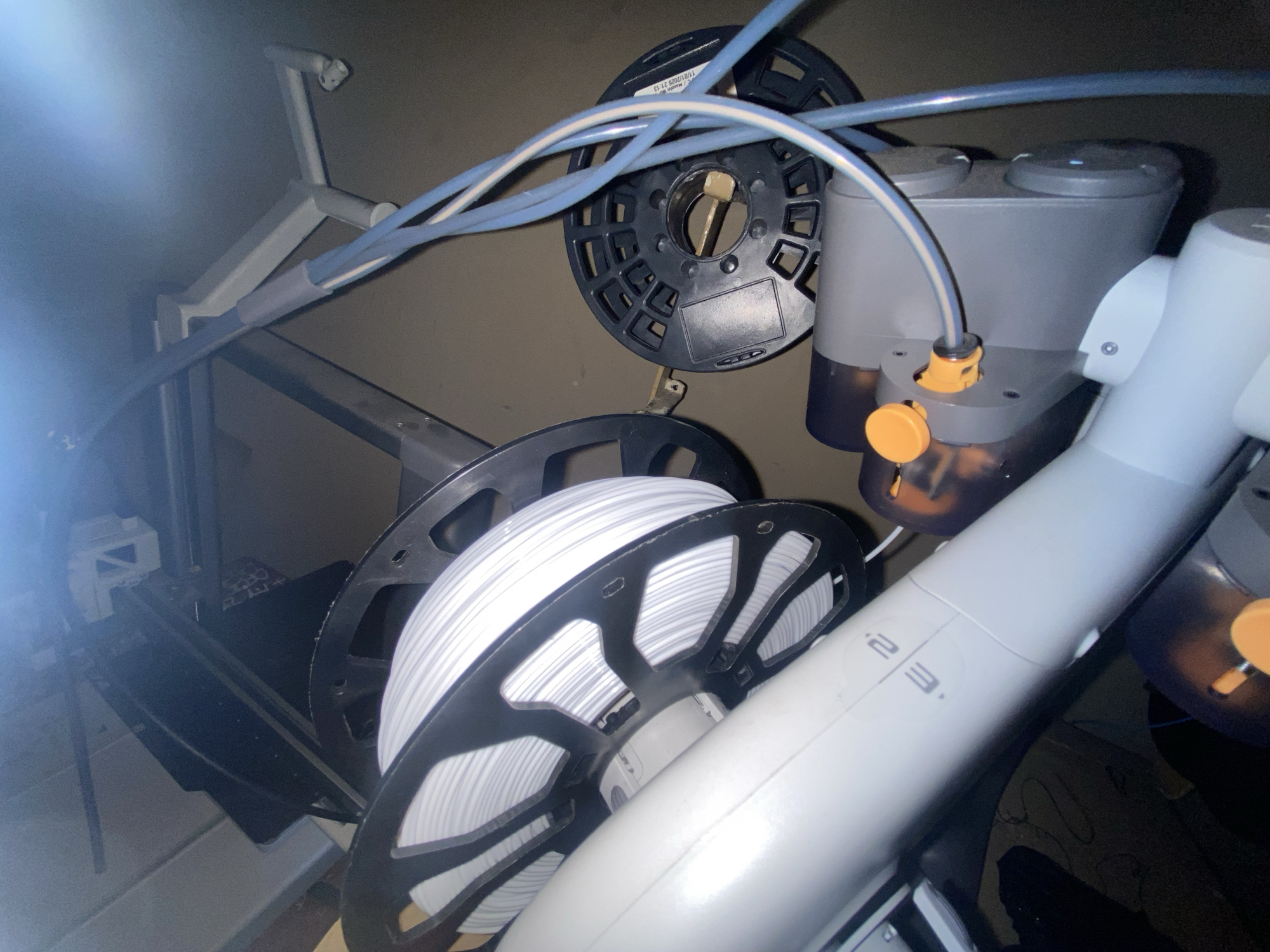 The image shows a 3D printer with two spools of filament. One spool contains white filament and the other contains black filament. Blue tubes run from the spools to the printer. A robotic arm labeled "2.3" is visible, connected to a device with orange knobs. The 3D printer appears to be located in an enclosed space, possibly a workshop or a laboratory. The location in Buenos Aires, Argentina, is unknown.