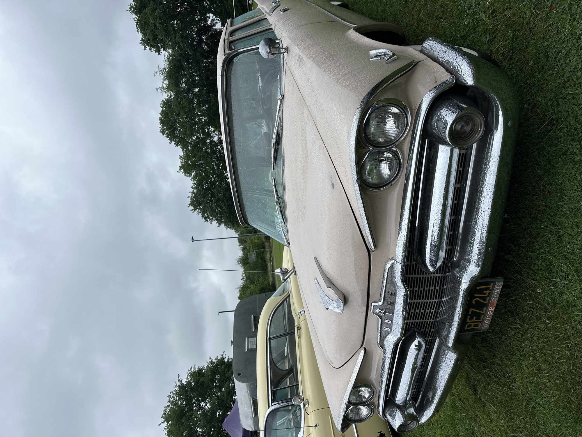 This image depicts a vintage car gathering in Deinste, Germany, under a heavily overcast sky. The primary subject is a light beige or pale pink 1950s Dodge sedan, positioned in the foreground and viewed from a low, slightly off-center front angle. The car's body, windshield, and chrome elements, including the grille, bumper, and headlight bezels housing dual circular headlights on each side, are covered in numerous small water droplets, suggesting recent rain or heavy dew. A prominent chrome hood ornament, resembling stylized wings, is centered on the hood. The grille features "DODGE" lettering within its central emblem. A black front license plate with yellow characters "BEZ 241" and "DODGE BOYS" text is mounted on the lower grille. A chrome side mirror is visible on the driver's side, and the windshield wipers are in their resting position.
Behind the main Dodge, to the left, the rear section of a yellow vintage coupe or sedan is partially visible, featuring a curved roofline and chrome trim. Further back, a distinctive silver-grey vintage travel trailer with a rounded aerodynamic shape and horizontal ribbing is parked. A purple canopy or tent is partially visible to the far left. The vehicles are situated on a verdant, well-maintained grassy field that appears damp. The background consists of a dense line of green trees under a uniformly grey and cloudy sky, with a tall light pole visible in the mid-ground. There are no individuals present in the scene.
