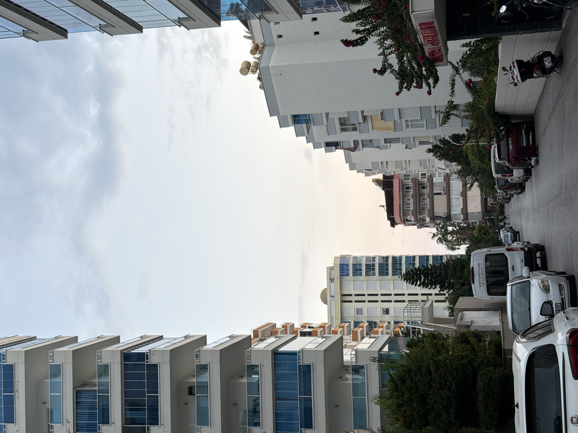 The image shows a street in a city with tall apartment buildings on both sides. The sky is overcast with some clouds. The buildings are modern, with white facades and balconies. On the left side of the street, there are several cars parked, including white cars and a white van. There are also trees and bushes along the sidewalk. On the right side of the street, there are more apartment buildings, some with visible solar panels on the rooftops. A scooter is parked in front of a building with a red sign. There are a few people visible walking in the distance. The overall impression is that of a residential area in a city, likely in a warm climate given the presence of flowering plants.