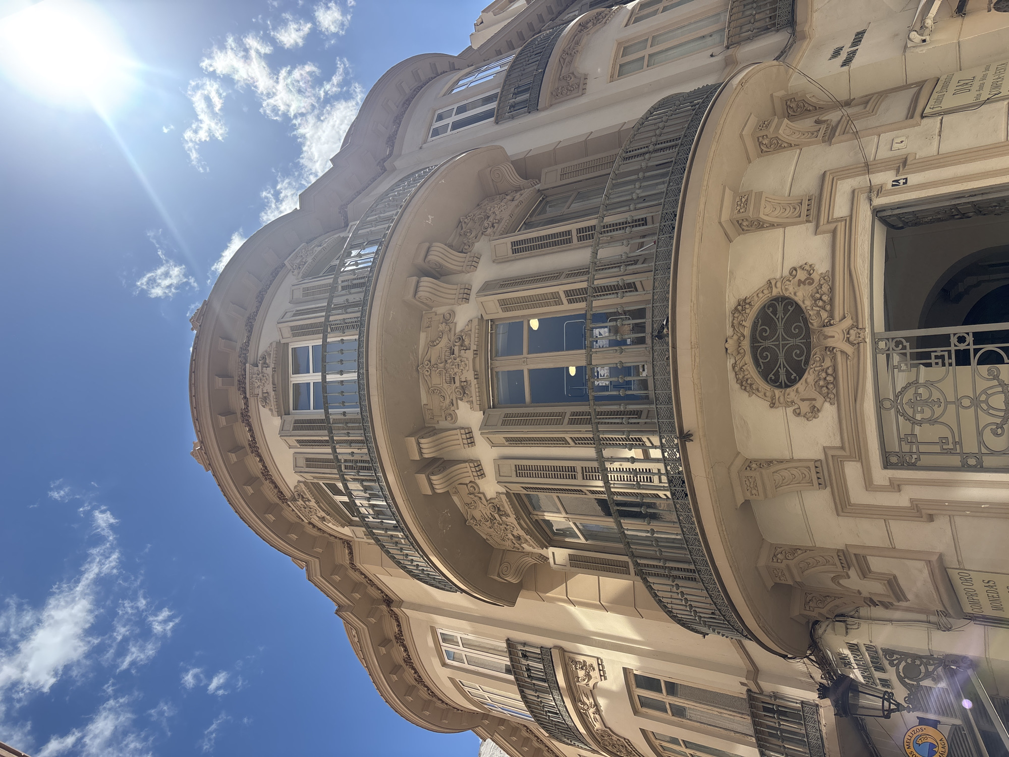 An upward-angled shot captures the facade of a cream-colored building in Málaga, Spain, under a blue sky dotted with scattered clouds and a bright sun. The architecture features curved balconies with wrought iron railings, and windows with louvered shutters. Ornate decorative elements are interspersed throughout the facade, including sculpted floral patterns and geometric shapes. A sign in Spanish hangs near the building's base. The building seems to be located in a historic or commercial area, evidenced by the architectural style and signage.