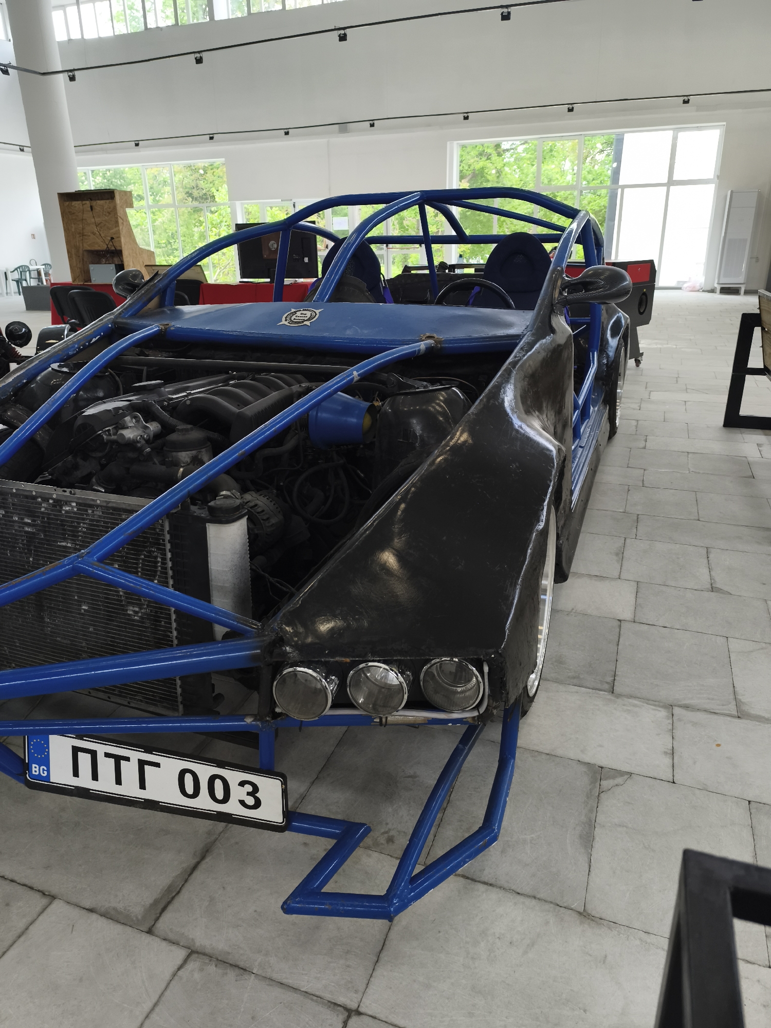 The image shows a custom-built, open-wheeled vehicle, likely a kit car or a heavily modified chassis. The vehicle has a blue roll cage and frame, with black body panels. The front of the car features three round headlights and a license plate reading "ПТГ 003" with the "BG" designation, indicating Bulgaria. The engine is visible, suggesting a front-engine configuration.
The vehicle is located indoors, in a space with tiled floors and large windows. Based on the license plate prefix "ПТГ," which corresponds to Targovishte, Bulgaria, it's likely the vehicle is located in an exhibition or display space within the city. Other objects in the surrounding area include chairs, tables, and cabinet-like furniture along the perimeter of the room. The lighting is bright and airy.