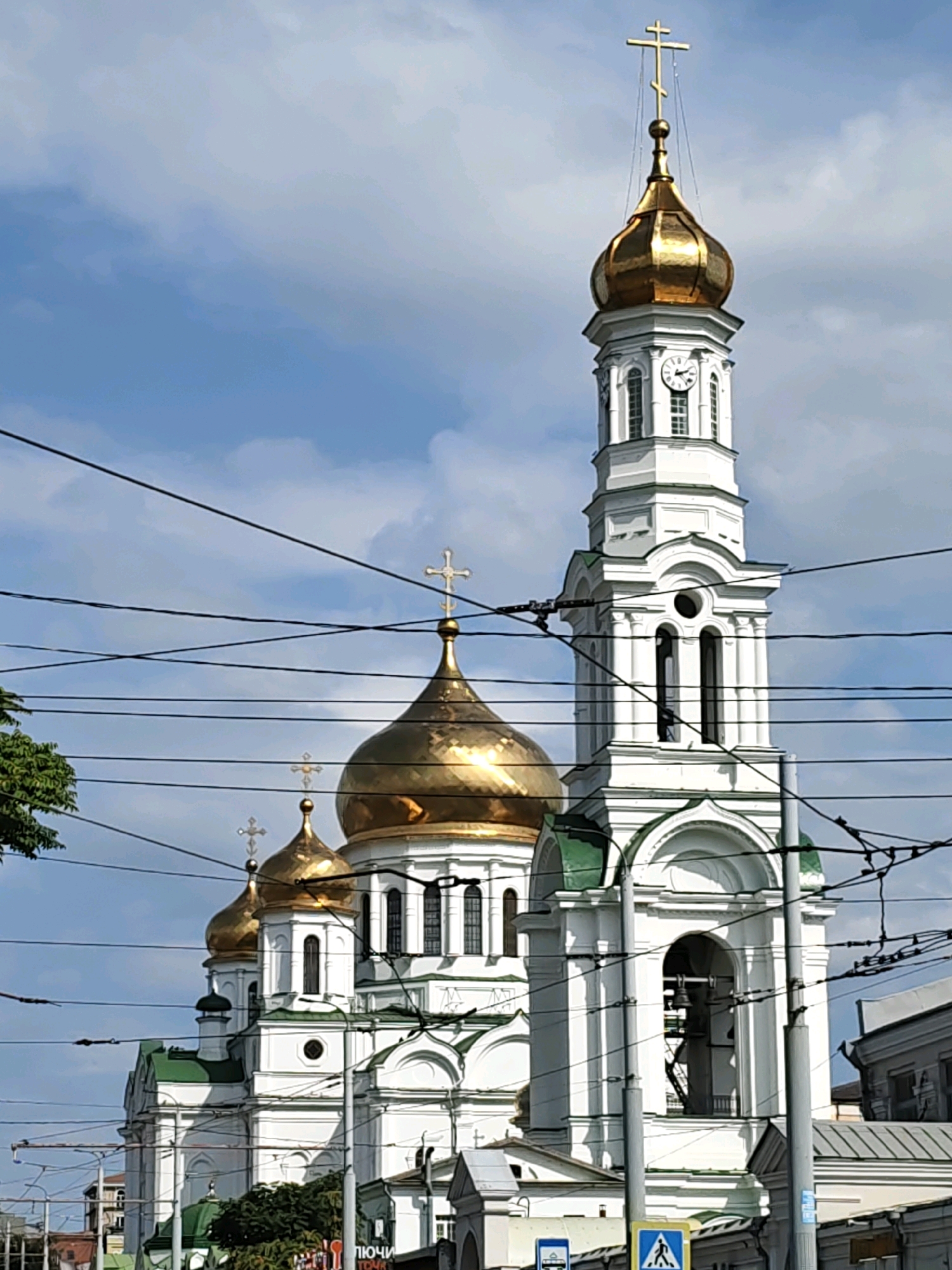 The image shows a white church with golden domes and a white bell tower, located in Rostov-on-Don, Russia. The church has multiple domes, each topped with a cross. The bell tower has a clock face and an arched opening where a bell is visible. The scene is framed by a network of overhead wires, likely for an electric trolleybus system, which are a common sight in Rostov-on-Don. There are no people or specific actions depicted in the image, but the presence of the wires suggests an urban setting. The sky is blue with scattered clouds.